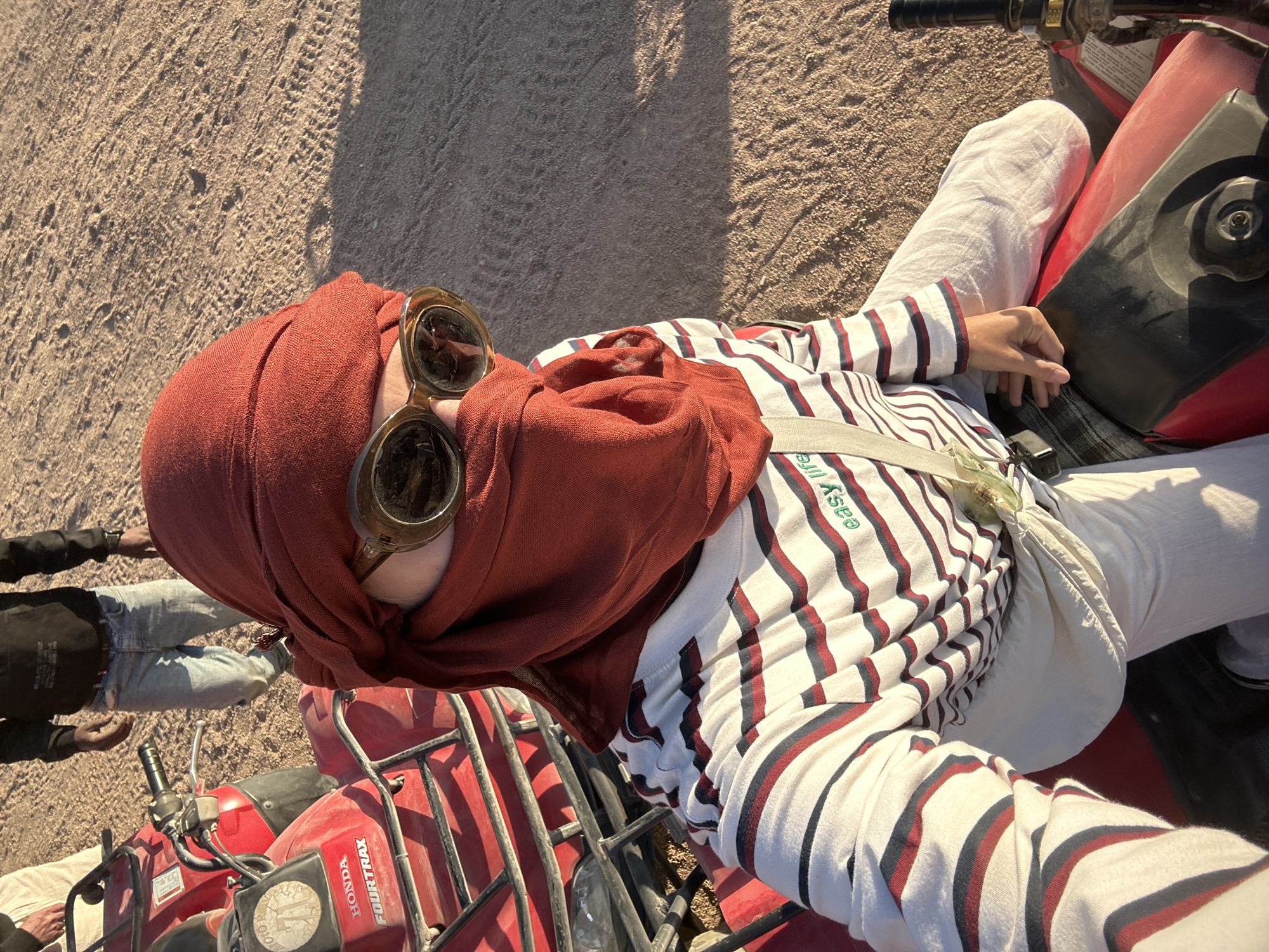 A person wearing a red headscarf and retro goggles sits astride a red Honda Fourtrax ATV in what appears to be a desert or sandy environment. The person is also wearing a striped shirt with the phrase "easy life" visible on the chest area, and white pants. Another person, partially visible, stands beside another ATV in the lower left. The ground appears to be sandy or dusty. Based on the details of clothing and environment, this could be in Sharm El Sheikh.