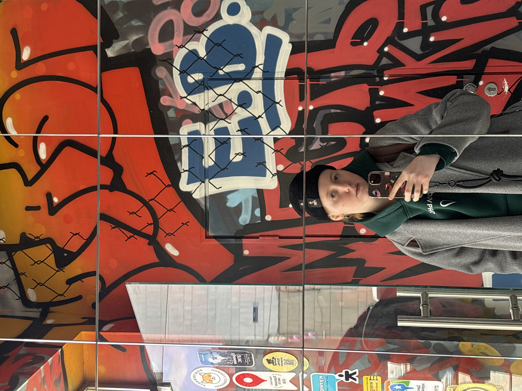 A young person is captured in a reflection on a window or glass panel, which is extensively covered in vibrant street art. The individual, dressed in a black beanie, a dark green hoodie, and a grey coat, is holding a smartphone up to take a selfie. Their face, looking towards the phone, shows a neutral expression.
The background of the reflection is a wall or surface adorned with bold graffiti, featuring large, stylized letters and shapes in bright red, orange, black, pink, and blue. A prominent blue and white piece resembles the characters 'SL' or '91', overlaid with a distinct black chain-link fence pattern that also appears across other sections of the graffiti. This fence pattern, with its sharp, uniform grid, could be painted onto the glass or part of the mural.
The reflective surface itself is divided by thin horizontal and vertical lines, suggesting window panes or a structural frame, which cut across the graffiti. In the lower left corner of the reflected view, beneath the main graffiti, various stickers are visible on what appears to be a separate surface. These stickers display a mix of graphics and text, with several phrases in Cyrillic, including "SLNC", "WECLO", "xobom66", "росавека-ские", "клубе", "НОЖНО", "THIENDIA", and "360", consistent with the location context of Perm, Russia. The person's green hoodie also has partially visible text, "DV PHO". The overall scene depicts a dynamic urban environment, blending personal interaction (selfie-taking) with public artistic expression.