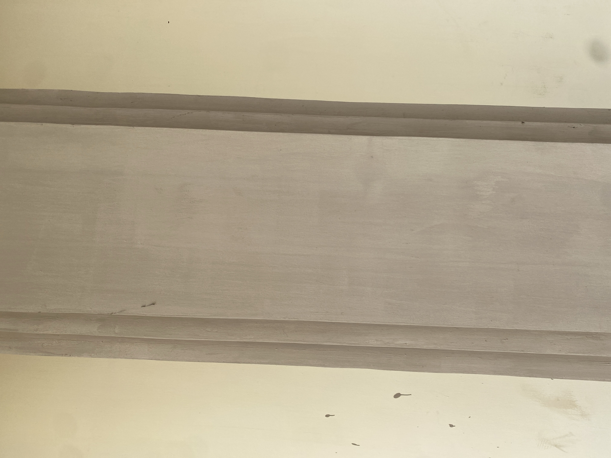 The image shows a close-up of a building's exterior wall with a vertical column. The wall is painted a light cream color. The column is made of concrete and has a textured finish. It is a neutral gray color and has three distinct vertical grooves, creating a fluted effect. There are some dark splatters on the cream-colored wall, resembling dirt or paint. The image appears to be taken during daylight hours, with natural light illuminating the scene. The location is Jalingo, Nigeria.