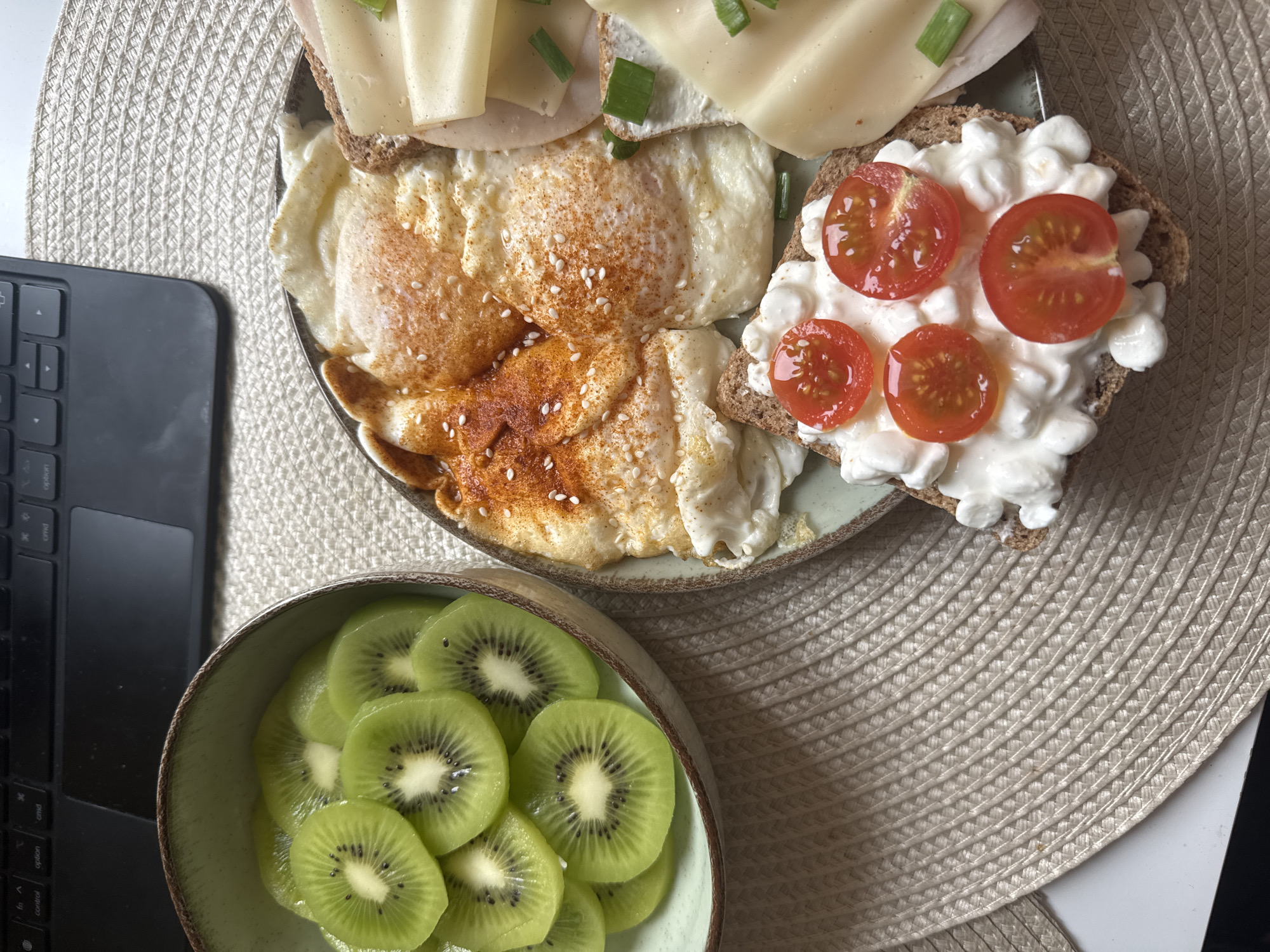 A top-down shot shows a round, woven beige placemat with a plate of food and a bowl of sliced kiwis next to a portion of a black laptop. The plate contains fried eggs, a slice of bread topped with cottage cheese and sliced cherry tomatoes, and another slice of bread with ham and cheese topped with chopped green onions. The kiwis are bright green and piled in a green bowl. The scene appears to be indoors, perhaps in an office or home workspace. There is no indication of the location being Sofia, Bulgaria.