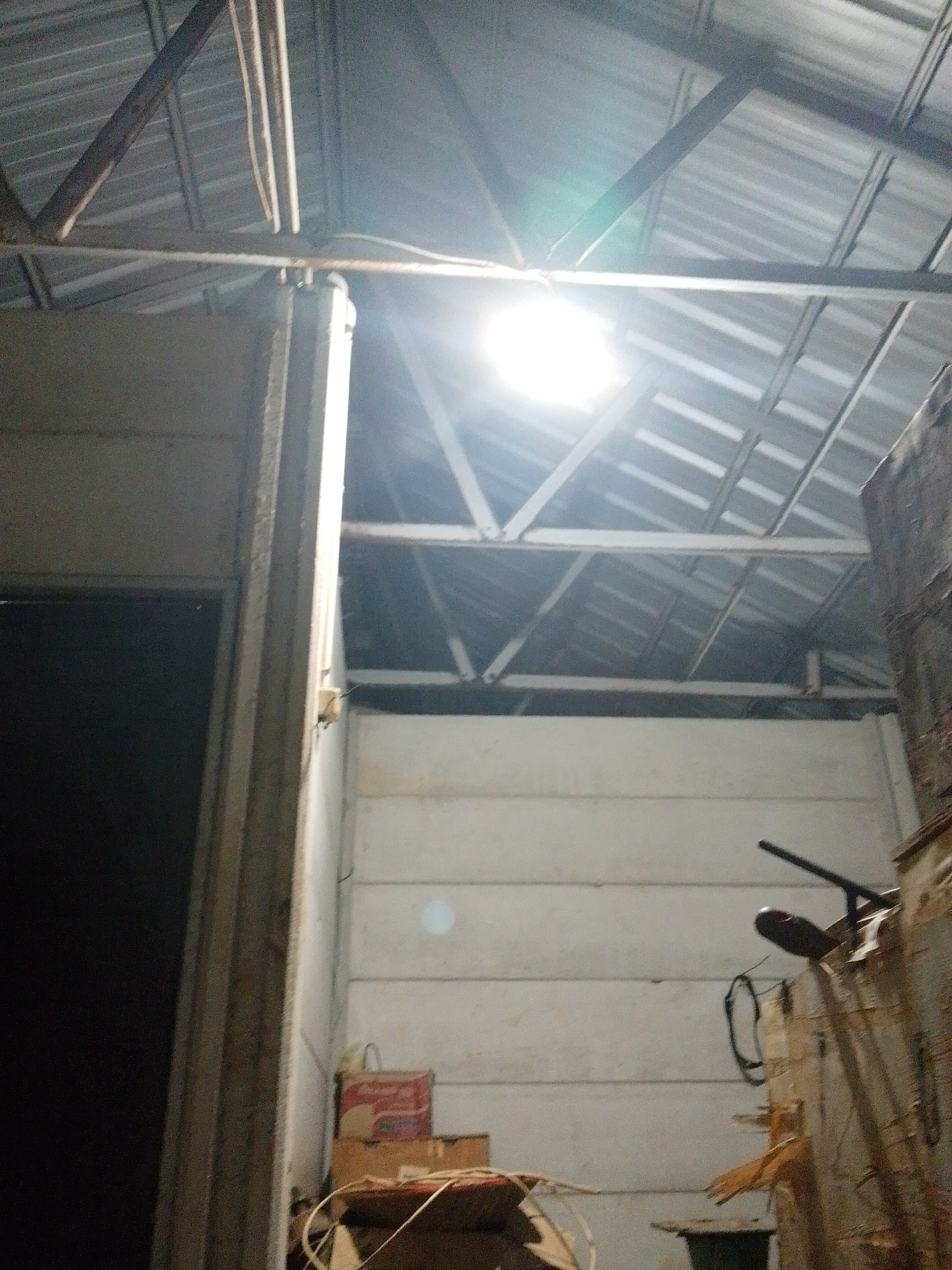 The image depicts an interior scene of what appears to be a partially constructed or storage area, with a metal roof supported by exposed beams. The lighting suggests it may be nighttime or dimly lit, with a bright light source in the upper center of the frame, possibly a bare bulb, creating a lens flare. To the left, a dark opening suggests a doorway or passage. The walls are constructed of horizontal panels. In the lower right quadrant, there are stacked boxes and debris, with a partially visible object that could be a tool or piece of equipment. A red and white box with a handle is prominent among the stacked items. The overall environment is utilitarian and appears somewhat disorganized. This scene is located in Bukit Ketok, Indonesia.