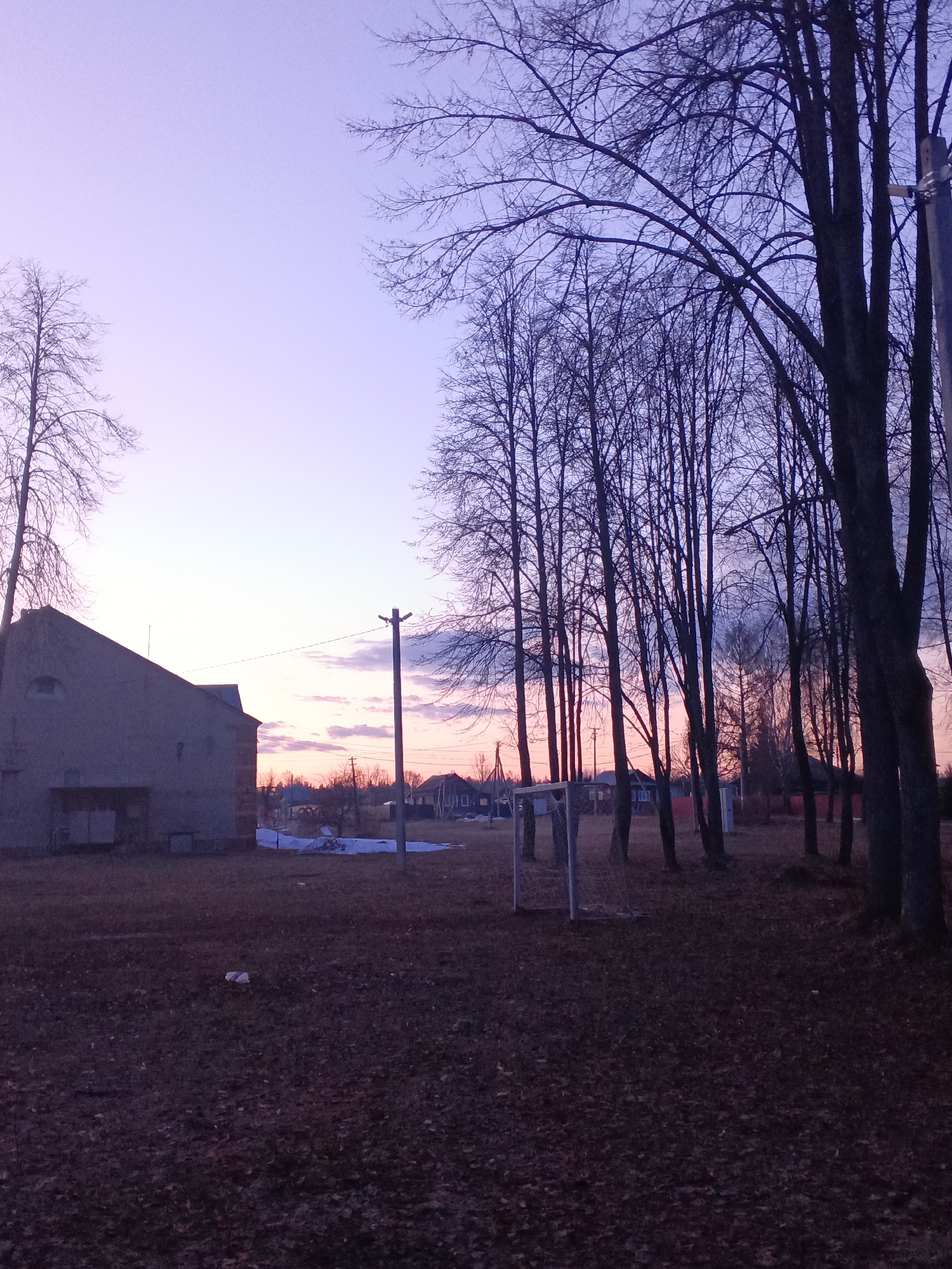 The image shows a dusky scene in Красный Маяк, Russia. The sky transitions from light purple to orange on the horizon. Silhouetted trees dominate the right side of the frame, while a building stands to the left. A utility pole is visible near the center. In the foreground, a soccer goal is situated on a field with fallen leaves. Patches of snow dot the landscape near the horizon. Houses can be seen in the distance.