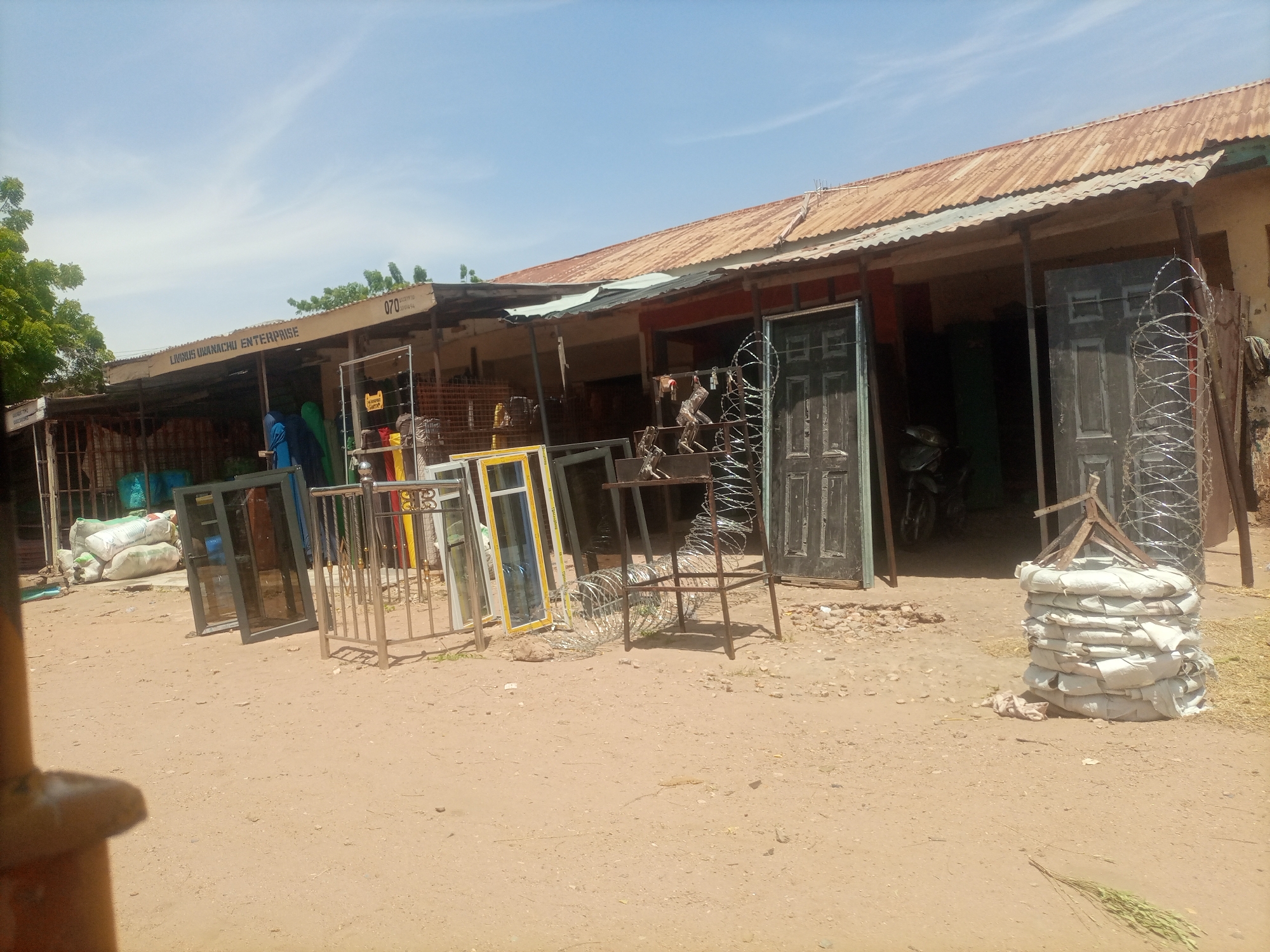 The image depicts an outdoor scene in the city of Arawa, Nigeria. In the foreground, a dusty, sandy ground is visible. To the left, a portion of a metallic structure, possibly part of a stall or building, is in the foreground.
The main focus of the image is a storefront or workshop area. Above the entrance, a sign reads "LIVINUS UBANACHU ENTERPRISE". The building has a corrugated metal roof that appears to be weathered. Various items are displayed and stored in front of the building, suggesting a business dealing in construction or building materials. These items include several window frames with glass, some appearing to be new and others possibly older. There are also metal gates or railings, and rolls of barbed wire. Two dark-colored doors are visible standing upright. In the background, stacks of white bags, possibly containing building materials like cement, are piled up. There are also some figures in the background, one appearing to be wearing blue clothing, and another person wearing dark blue and black. A motorcycle is partially visible inside the workshop. The overall environment suggests a commercial area in a town or city, with the building and displayed goods typical of such a location. The sky above is blue with some scattered white clouds.