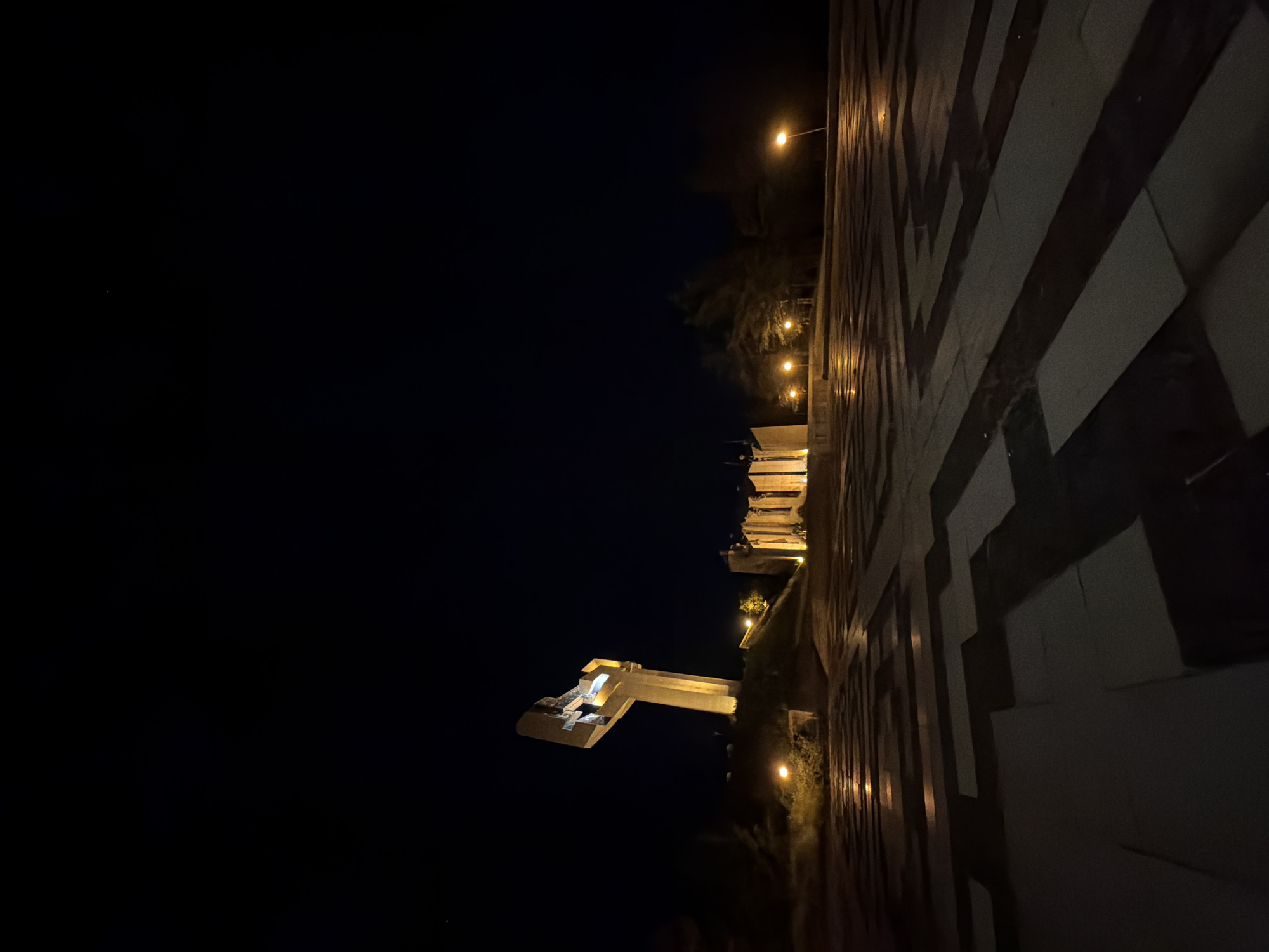 The image captures the Monument of the Bulgarian Communist Party in Stara Zagora, Bulgaria, at night. The monument is partially illuminated and contrasts with the dark night sky. Adjacent to the monument, there is a wall with a geometric pattern, also illuminated. Streetlights dot the foreground. No people are visible.