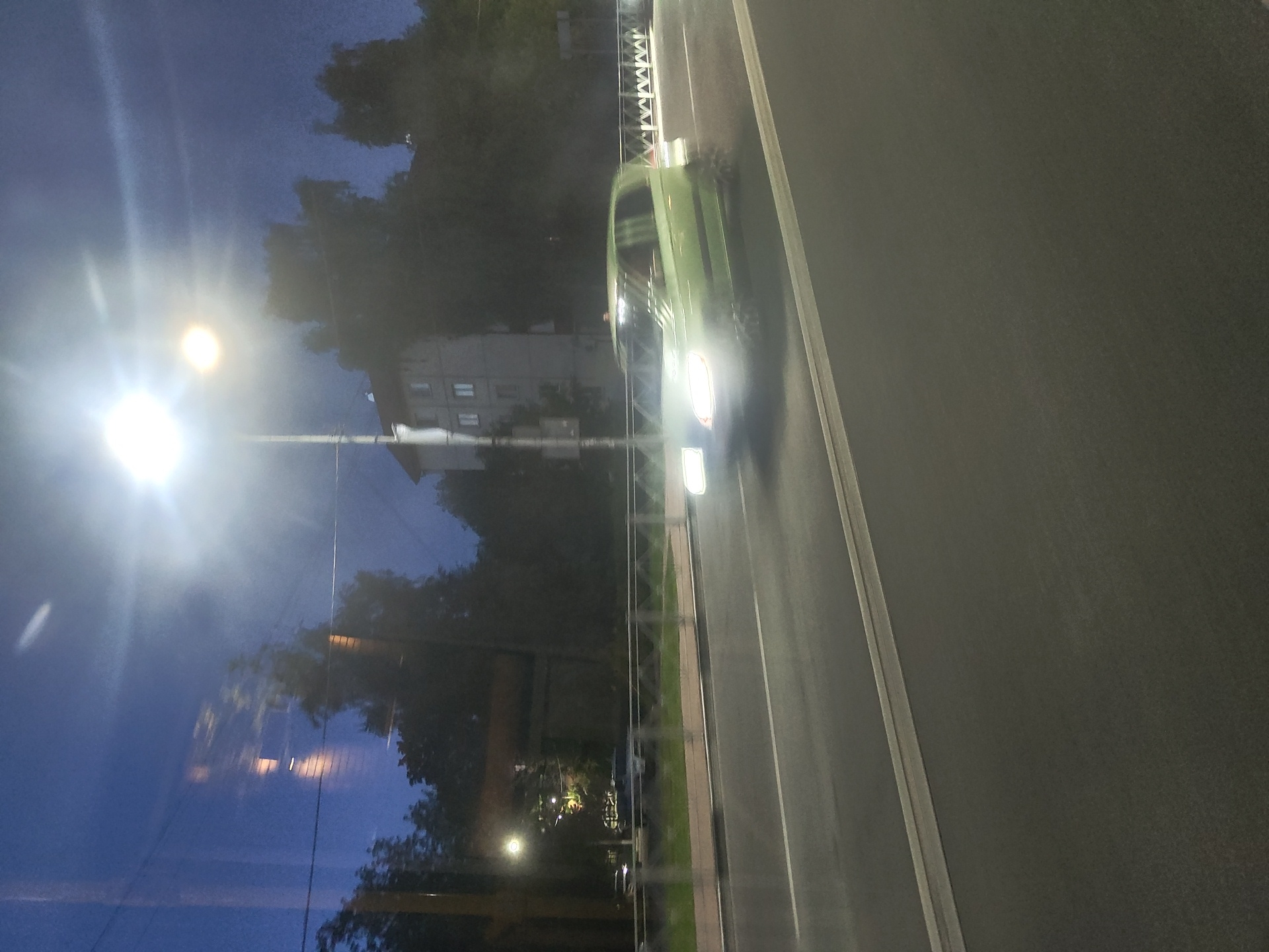 This image captures a motion-blurred view of a street scene at dusk or night in Ramenskoye, Russia. A light green car is driving on the road, its headlights brightly illuminated. The car appears to be a hatchback. A street lamp with a bright light source is visible to the left of the frame, casting a strong glow and contributing to the overall blur. In the background, there is a multi-story building with visible windows and trees silhouetted against the dark blue sky. A metal fence with a decorative pattern runs along the side of the road. The road itself is paved and has white lane markings. Reflections are visible on what appears to be a window, suggesting the photo was taken from inside a vehicle or building.