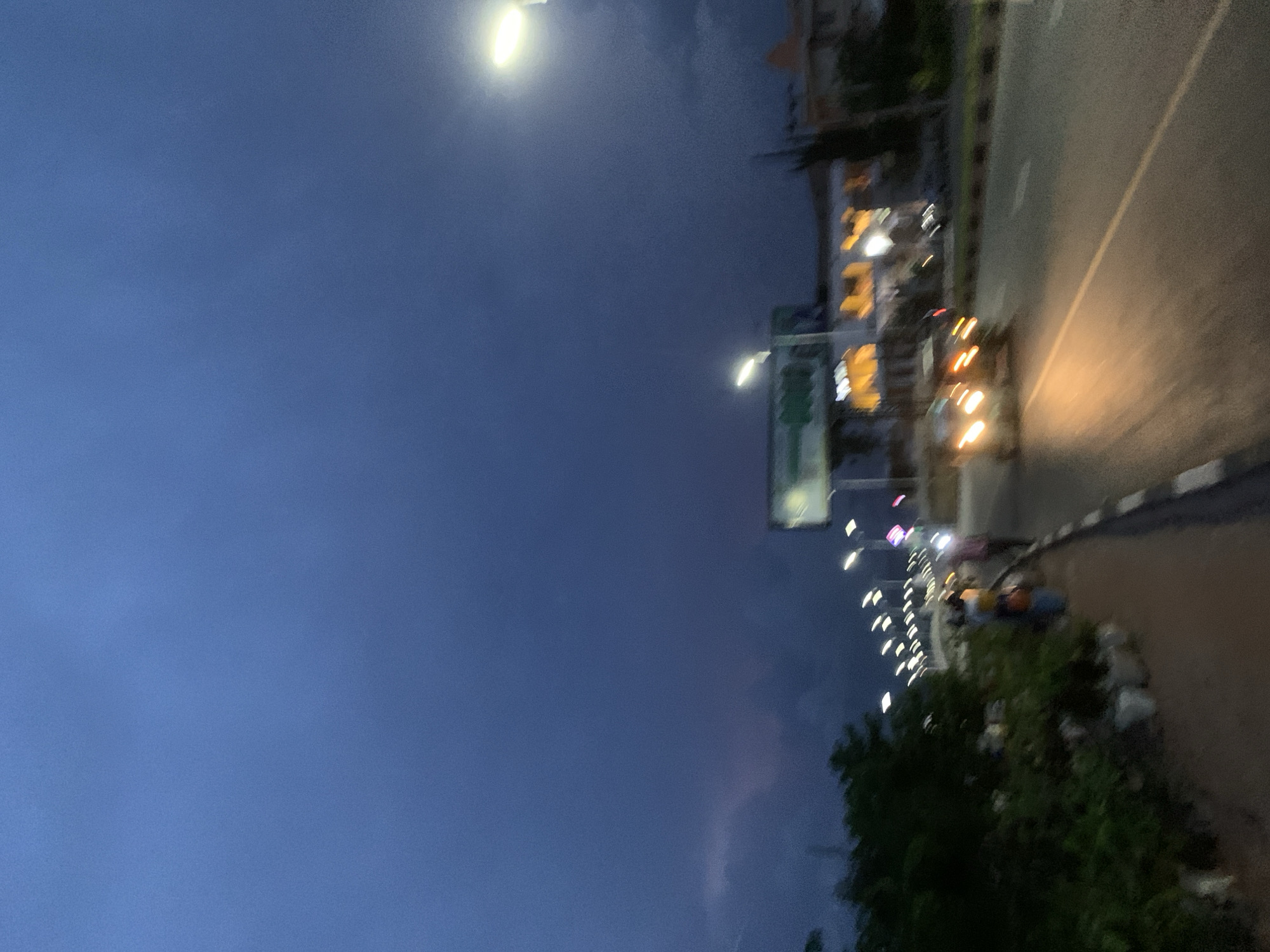 The image depicts a street scene at dusk or dawn in Jos, Nigeria. The sky is a deep, moody blue, suggesting an impending storm or the aftermath of one, with hints of purple and pink in the clouds. A highway runs through the scene, with a car's headlights brightly illuminating the asphalt. The car appears to be traveling at speed, creating motion blur.
On the left side of the road, a sidewalk is visible, bordered by lush green foliage and some white-capped structures, possibly planters or waste bins. A few individuals are present on the sidewalk, their figures blurred due to the low light and motion. One person in the foreground is wearing a yellow and white top. Further down the street, a row of streetlights creates a repeating pattern of illumination against the darkening sky.
Buildings line the opposite side of the highway, their windows glowing with artificial light, indicating activity within. A large billboard with a green sign is also visible, partially obscured by a streetlight. The overall impression is one of a busy urban environment transitioning from day to night, with the weather adding a dramatic element to the scene. The motion blur suggests the photograph was taken from a moving vehicle or with a slower shutter speed.