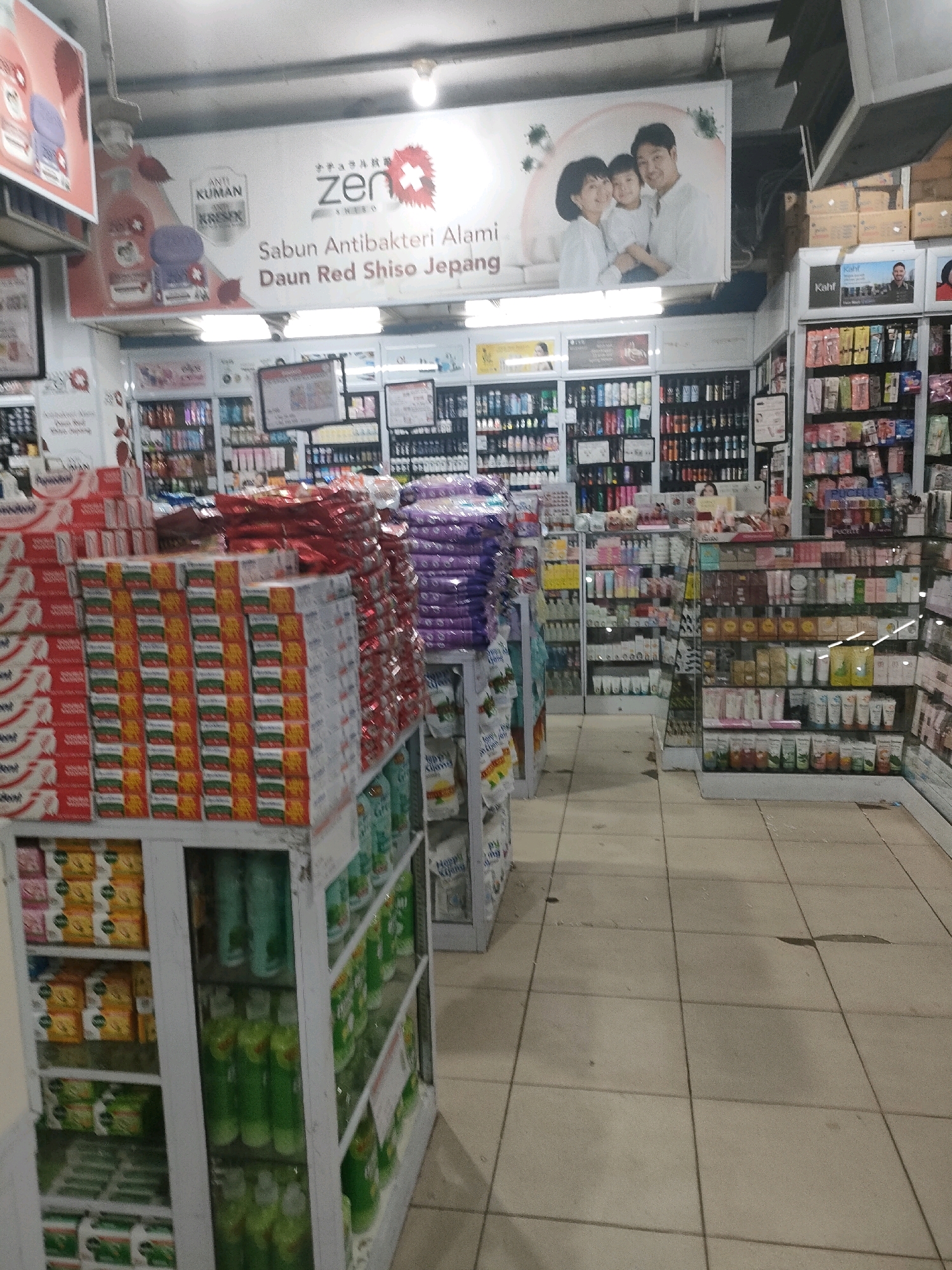 The image depicts the interior of a retail store, likely a supermarket or a drugstore, with aisles filled with various products. In the foreground, there are shelves stacked with packaged goods, including boxes of what appears to be soap or detergent, and some larger, colorful bags. To the right, more shelves are visible, stocked with rows of bottles and containers, some of which are translucent and contain green liquid, suggesting cleaning supplies or personal care items. Further into the store, additional aisles are lined with shelves showcasing a wide variety of products, including skincare items, cosmetics, and other household goods. A large promotional banner is visible hanging from the ceiling, featuring an image of a family and product branding for "Zen" soap, along with text that mentions "Sabun Antibakteri Alami Daun Red Shiso Jepang." The flooring appears to be tiled, and overhead lighting illuminates the space. There are no people visible in the frame.