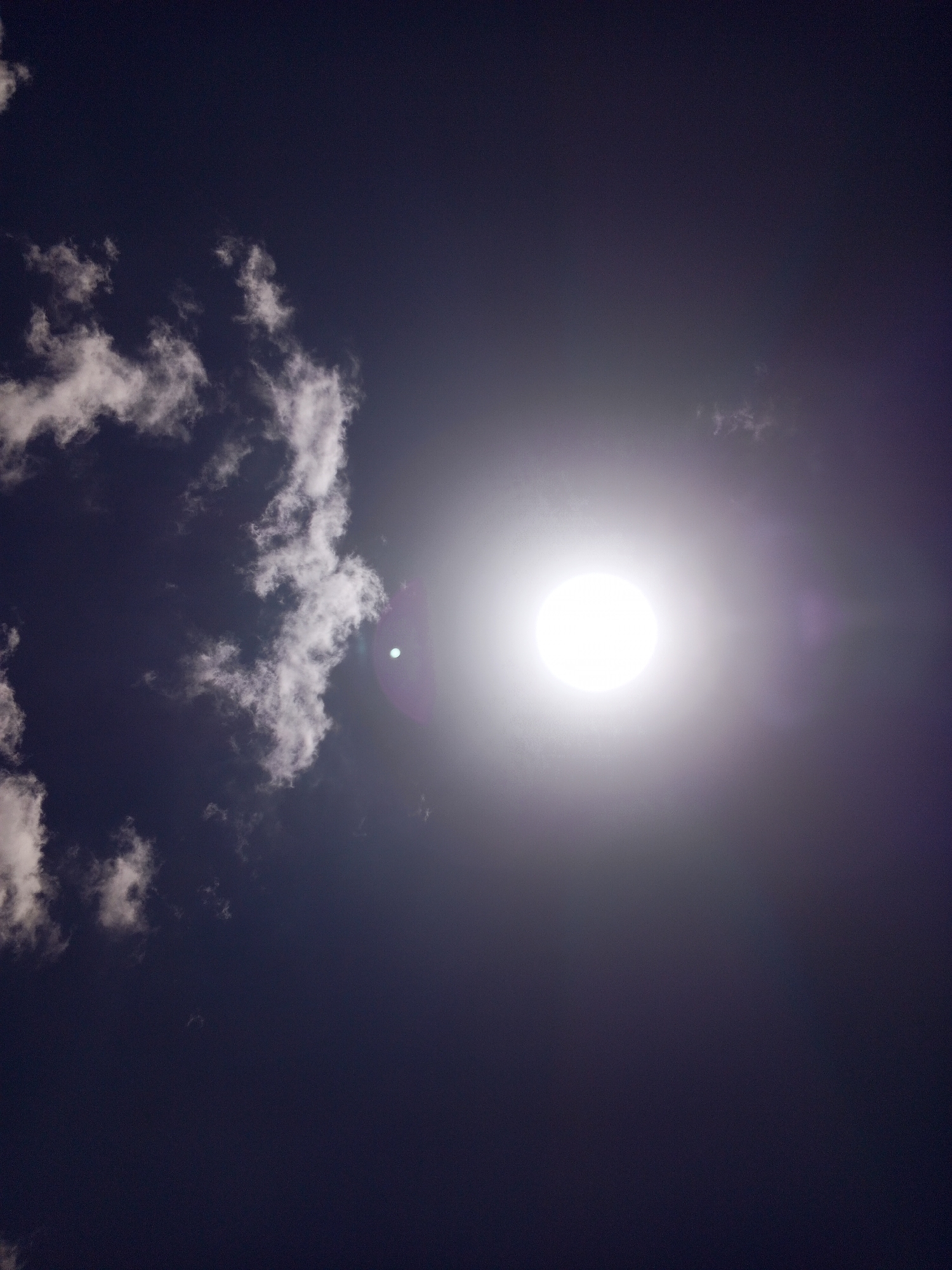 A view of the sky shows scattered white clouds in the upper left quadrant against a dark blue-violet backdrop. A bright, round light source, potentially the sun, occupies the center of the frame, emitting a strong glare and a faint lens flare towards the right. The scene suggests daytime lighting. No people or identifiable objects are visible besides the sky and clouds. The location is assumed to be Altagracia, Venezuela, per the prompt's request for location inclusion.