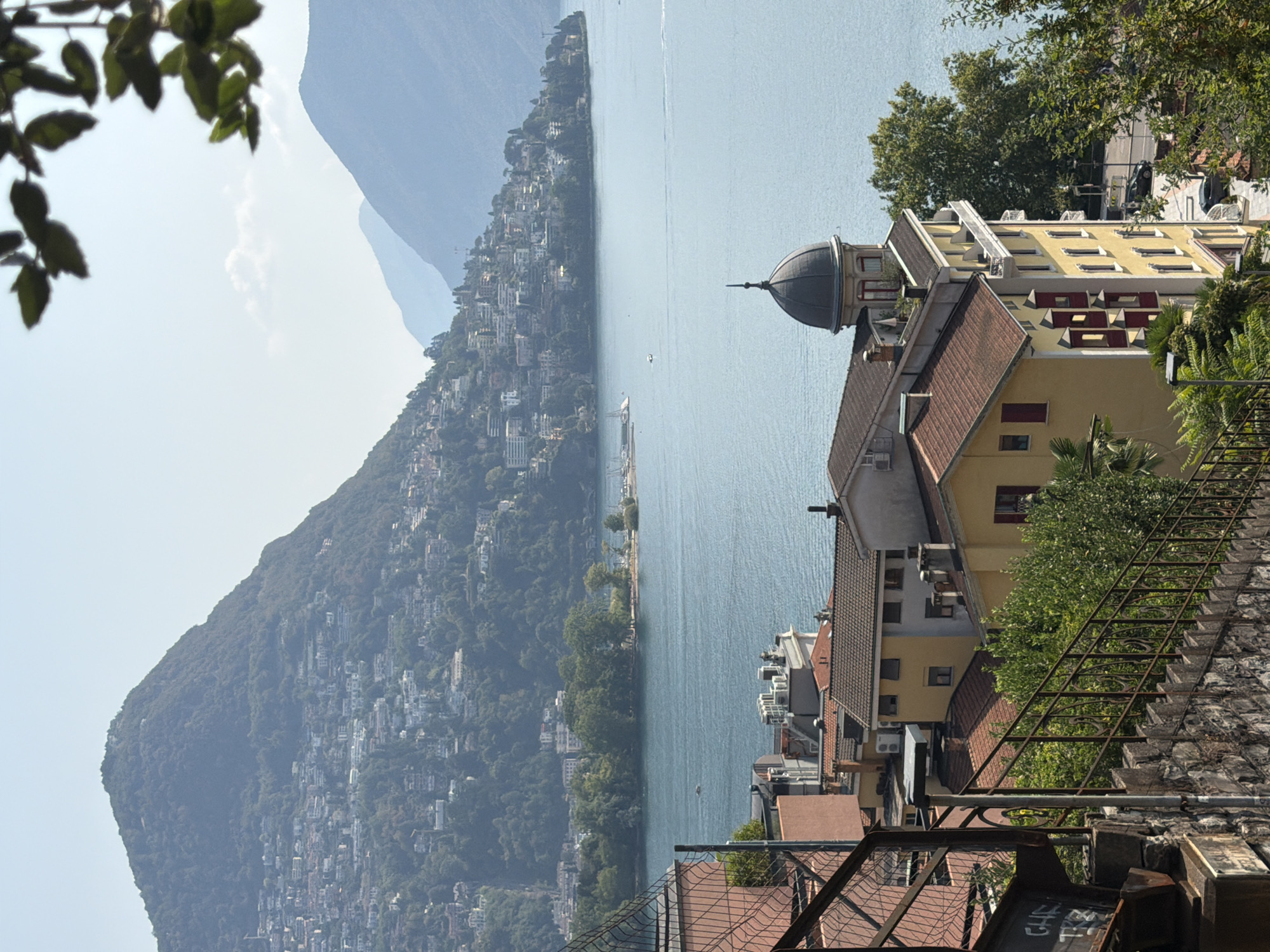 This image captures a scenic view of Lugano, Switzerland, overlooking the serene Lake Lugano and the surrounding verdant mountains. In the foreground, a stone staircase with a rusty metal railing leads the eye upwards. To the left, part of a red metal fence and a concrete structure with graffiti are visible. The middle ground features a cluster of buildings with red-tiled roofs, predominantly yellow and beige facades, some with multiple stories and balconies. One building is adorned with a distinctive domed turret. Green foliage and trees are interspersed among the structures and climb the mountainside. Across the lake, the slopes of the mountains are densely populated with houses and buildings, forming a picturesque hillside cityscape. The water of the lake is a clear blue, with a few small boats visible in the distance. The sky is a pale blue with a few wispy clouds, and sunlight illuminates the scene, casting soft shadows. The overall atmosphere is tranquil and idyllic, showcasing the beauty of Lugano's natural and urban landscape.