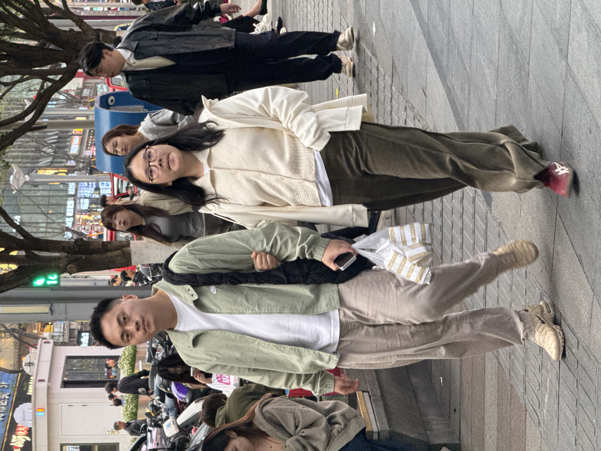 A group of people is walking along a street in Liangjiang, China. A woman in glasses is wearing a white cardigan, a white t-shirt, and green pants, and walking on a gray bricked sidewalk. A man in a green jacket, a white shirt, and tan pants, has a black bag, is also walking on the sidewalk. In the background are scooters, a building with Chinese lettering on it, and trees.