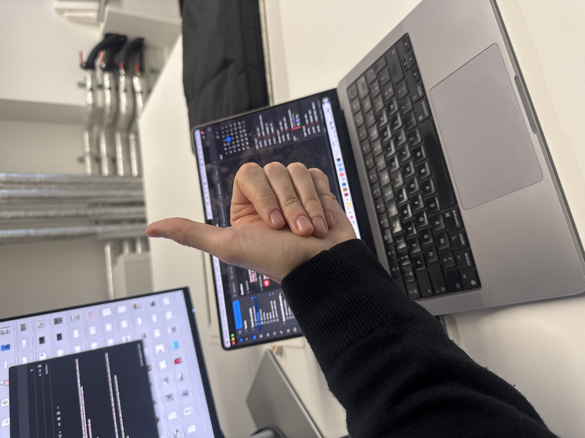 Here is a detailed description of the image:
**Scene Overview:**
The image is an overhead shot of a desk or workstation setup, likely indoors. The primary focus is on a person's hand giving a thumbs-up gesture, positioned in front of a laptop and an external monitor.
**Subjects and People:**
* **Hand:** The main subject is a hand giving a thumbs-up. The hand appears to be fair-skinned with short fingernails. It is attached to an arm wearing a black, long-sleeved sweater.
**Objects:**
* **Laptop:** A silver-colored laptop is partially visible on the right side of the frame. The keyboard and trackpad are visible.
* **External Monitor:** To the left, an external monitor displays what seems to be code or data.
* **Additional Display:** A second, smaller display is visible below the first monitor, also showing code or data.
* **Pipes:** A series of pipes run vertically against the wall in the background.
**Environment:**
* **Indoors:** The presence of a desk, monitors, and internal fixtures indicates an indoor setting.
* **Modern Office or Workspace:** The equipment suggests a professional workspace environment, possibly an office or a tech-related setting.
**Actions and Interactions:**
* **Thumbs-up:** The hand is giving a thumbs-up, suggesting approval, agreement, or a positive sentiment.
**Additional Details:**
* The overall lighting is somewhat bright and diffused, indicating a well-lit interior.
* The arrangement of the devices suggests a workspace designed for productivity, such as software development or data analysis.
There is no information to infer that this is in Paris, France.