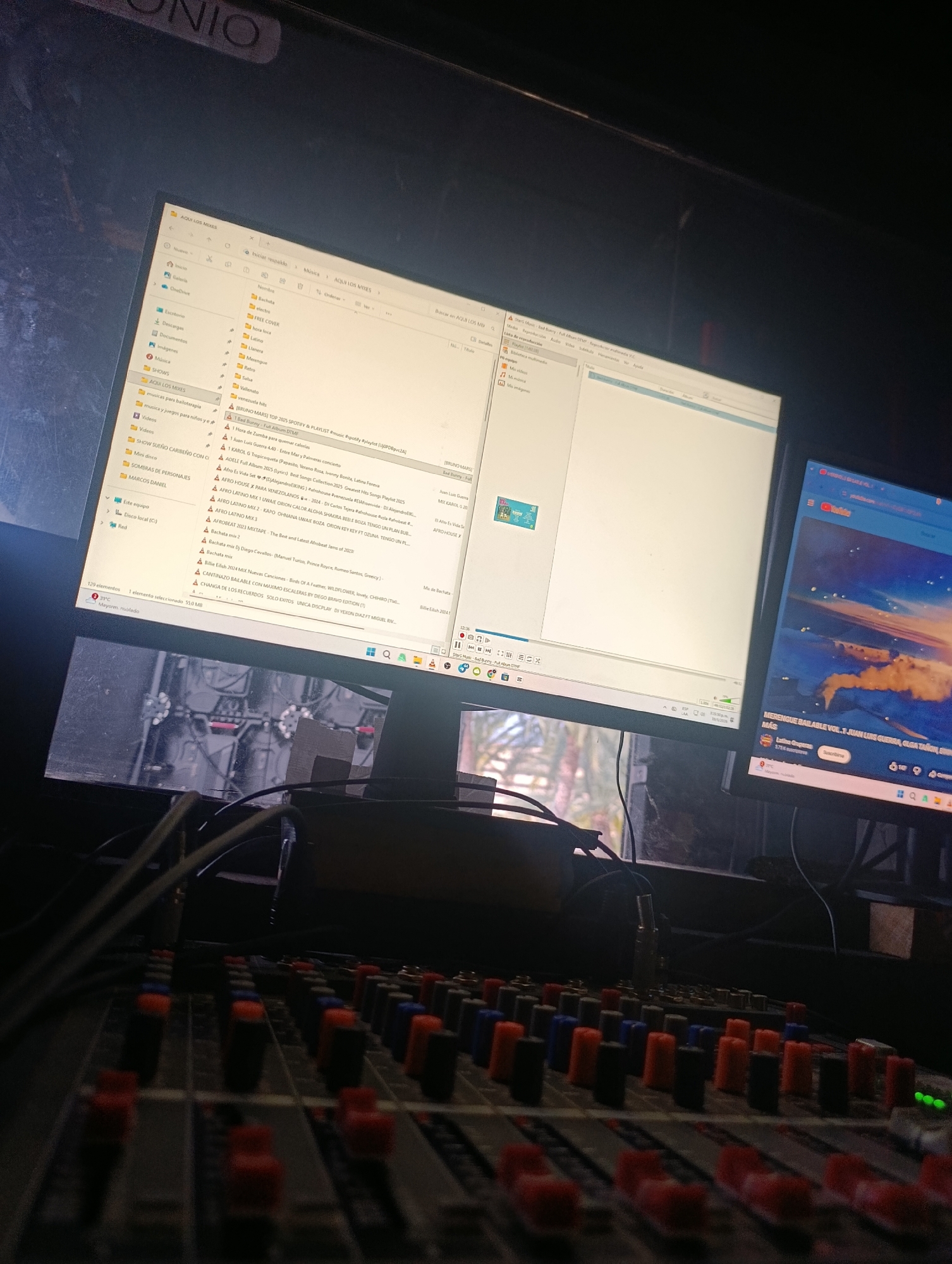The image shows a dimly lit recording or DJ setup, likely indoors, with audio equipment in the foreground. A mixing console with various knobs and sliders dominates the lower portion of the frame.
Behind the console, two computer monitors are visible. The left monitor displays a file directory with folders such as "AQUI LOS MIXES" and files like "Bachata," "Electro," and various music tracks, including artists like Bruno Mars, Bad Bunny, and Karol G. The right monitor is displaying a YouTube video with the title "MERENGUE BAILABLE VOL.1 JUAN LUIS GUERRA, OLGA TAÑON..."
The location is identified within the file directory visible on the left monitor, which shows a folder named "AQUI LOS MIXES." This matches a city location in Porlamar, Venezuela, where the user also identified it.