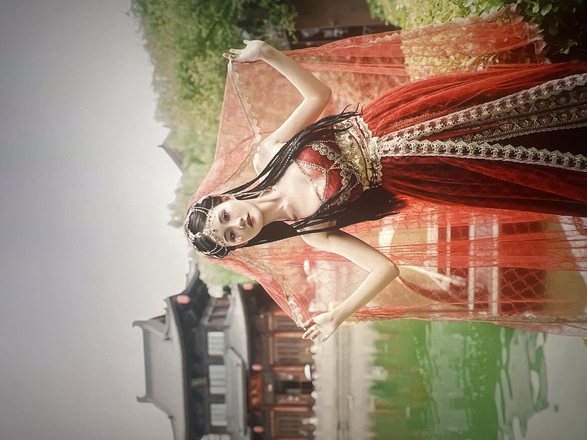 A young woman with dark braided hair adorned with a jeweled headband and facial glitter poses outdoors, dressed in a elaborate red and gold costume. Her dress features intricate gold embroidery and layered red fabric, with a sheer red veil draped over her head and shoulders. She is positioned in front of a traditional Chinese-style building, likely in Liandu District, China. The building has dark wooden elements and distinctive rooflines, and it overlooks a body of green water. The scene appears to be set during the daytime, with soft lighting suggesting overcast weather or shade from surrounding greenery. The woman's expression is serene and contemplative.