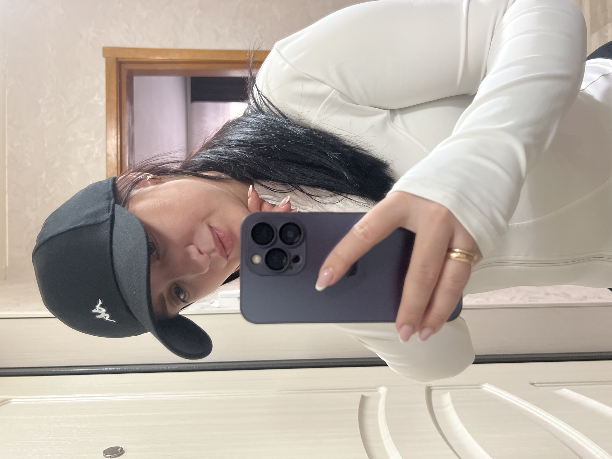 An indoor mirror selfie captures a young woman with dark, flowing hair, wearing a white long-sleeved top and a black baseball cap. The cap features a white embroidered logo, which appears to be a stylized "Kappa" emblem with two figures. She holds a dark-colored smartphone, equipped with a prominent triple-camera array, in her right hand, using it to capture her reflection. Her face is partially visible, showing her left eye, nose, and a slightly smiling mouth. Her right hand displays light-colored fingernails with French tips, and a gold ring is worn on her ring finger.
The background behind her head features a light-colored, textured wall. Above her shoulder, a wooden door frame is visible, leading into another room with lighter walls. The foreground reveals a reflective, light-colored surface, possibly a floor or another large panel, which shows architectural details like grooves or raised sections, and the reflection of her white top. A small, metallic, circular object rests on this surface in the bottom center. The woman is actively engaged in taking the photograph, indicated by her posture and the phone's position. The setting suggests a private or domestic interior, located in Марковский, Russia. The time of day is not discernible, and no specific emotions are strongly expressed beyond a neutral or slightly pleasant demeanor.