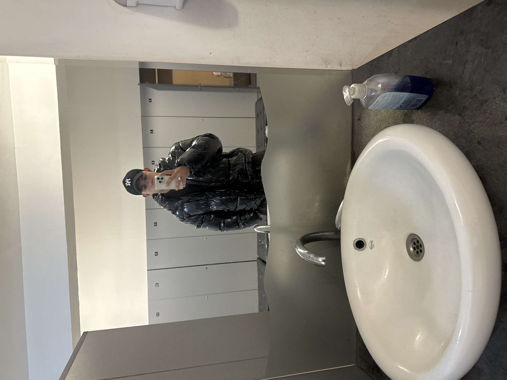 The image shows a person reflected in a bathroom mirror. The person is wearing a black puffy jacket and a baseball cap and is holding a phone up to take a photo. Behind the person in the mirror are a series of lockers. Next to the mirror is a white sink with a faucet and a bottle of soap. The bathroom has white walls and a grey floor. The location of this image is not discernible from the image alone. It is not possible to determine if it is in the city of Panagyurishte, Bulgaria.