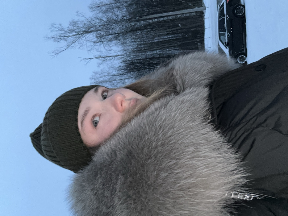 A person wearing a green beanie and a fur-lined jacket stands outdoors in a snowy environment. Bare trees are visible in the background, along with a dark-colored vehicle parked near a snow-covered area. The jacket has a large, gray fur collar. The person's face is partially visible, showing light skin and light-colored eyes.