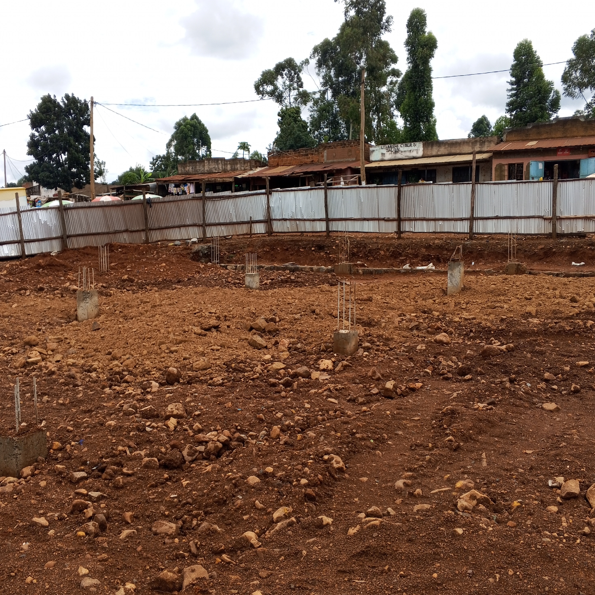 This media file captures a construction site under an overcast sky, likely in a semi-urban or rural area of Borabu Chitago ward, Kenya. The foreground and midground consist of a large, open plot of reddish-brown, uneven earth scattered with numerous rocks and small debris. Several concrete footings with protruding rebar reinforcements are visible across the site, indicating the foundational stage of a new building project.
In the background, a temporary fence constructed from corrugated metal sheets supported by wooden posts delineates the perimeter of the site. Beyond this fence, a row of simple, single-story buildings with corrugated iron roofs can be seen. These structures appear to be local shops or residences. Prominently displayed on one of these buildings is the text "UTUMISHI CYBER & SALON," suggesting the types of businesses operating nearby. Tall, leafy trees are scattered behind and around these buildings, reaching towards the light grey, cloudy sky. No people are visible in the image. The scene depicts a quiet moment in the early development phase of construction.