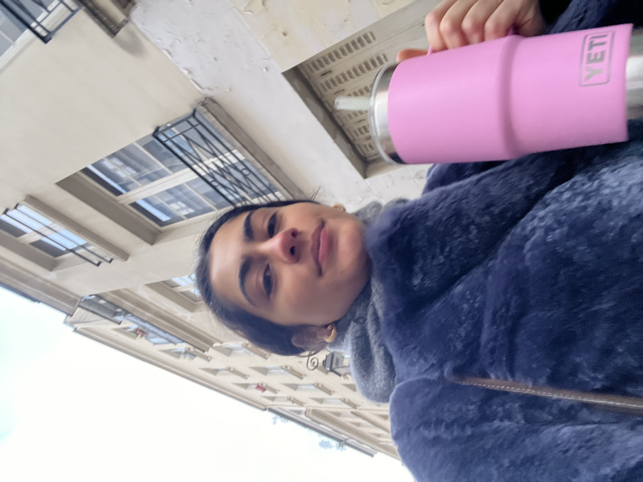 An upward-angled outdoor photograph depicts a young adult female from the chest up, positioned on a street in Paris, France. She has dark hair pulled back and wears a navy blue faux fur coat over a grey hooded garment, with gold hoop earrings visible. In her right hand, she holds a large, pink YETI tumbler with a silver base and lid, and a white straw. The YETI logo is clearly visible on the tumbler. The background features multi-story, cream-colored Haussmannian-style buildings with multiple windows, some equipped with narrow black wrought-iron balconettes. A historic street lamp fixture is attached to the building on the left. The sky is bright and overcast.