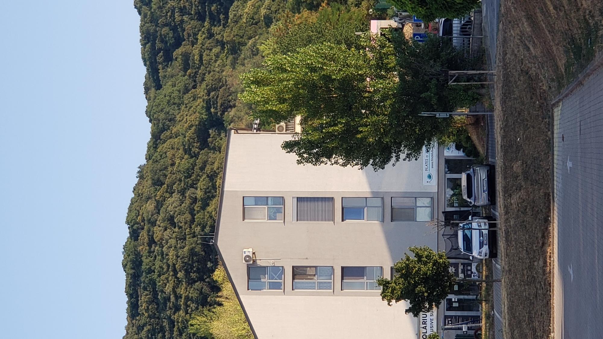 The image shows a modern apartment building with a light grey facade, featuring multiple windows and balconies. On the ground floor, there are commercial spaces with storefronts. Two cars are parked in front of the building: a white SUV and a silver hatchback. Behind the building, a dense, green forest covers a hillside under a clear blue sky. The scene appears to be in a suburban or semi-urban area of Bratislava, Slovakia, likely in a district with proximity to natural landscapes. The time of day appears to be late afternoon or early evening, with the sun casting shadows.