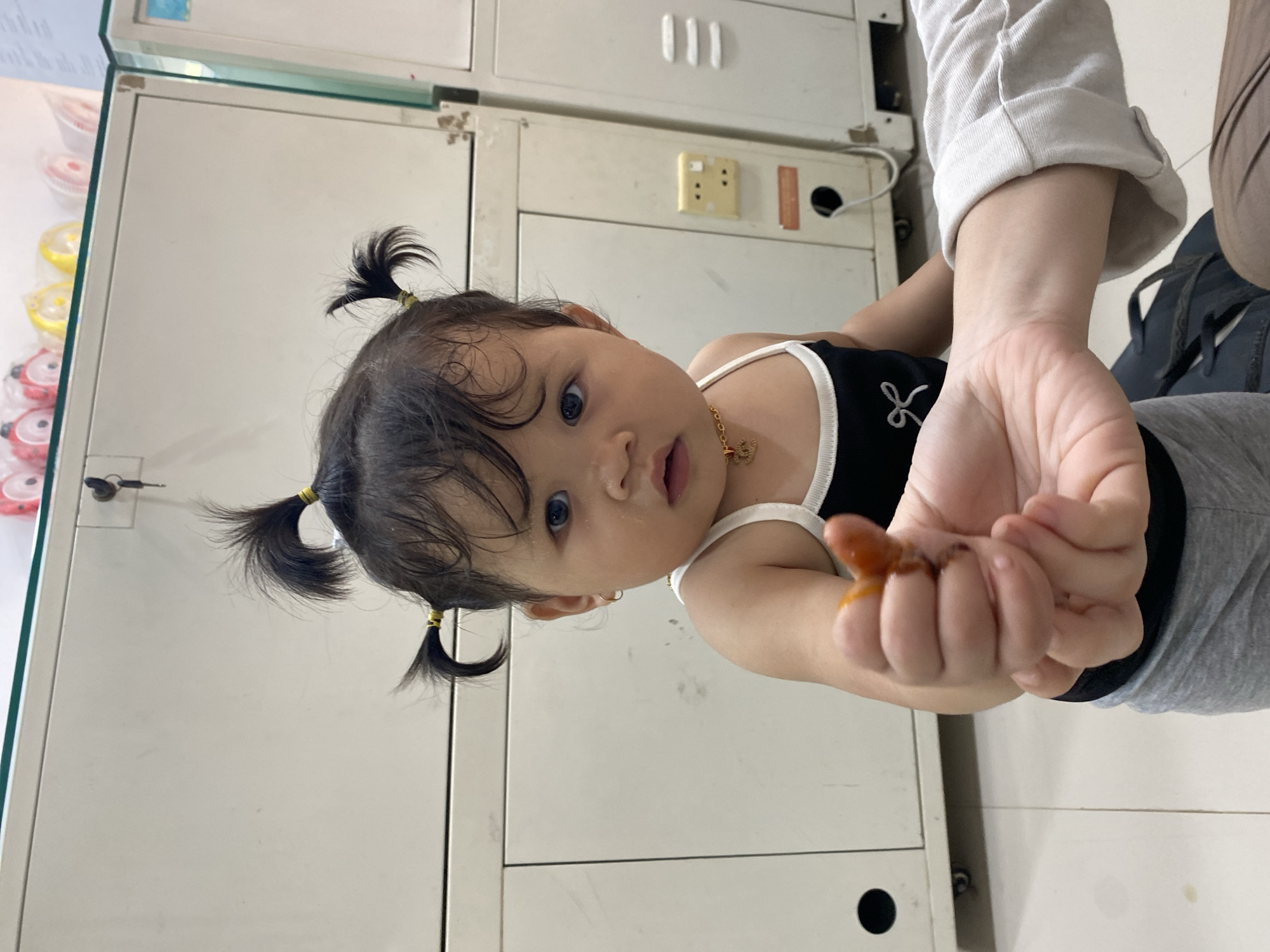 A young child with dark hair in two pigtails is the subject of this close-up, eye-level shot. The child is wearing a black tank top with white trim and a gold necklace. They are holding a small, orange object in their left hand, which appears to be food. The child's expression is neutral, with wide eyes looking directly at the camera. Another hand, presumably an adult's, is visible in the foreground, reaching towards the child. The background consists of a white, cabinet-like structure with some electrical outlets and ventilation grates. The lighting is bright and appears to be artificial. The scene is indoors, likely a home or a public facility, given the presence of the cabinets and electrical fixtures. No specific details suggest a location in Ta', Indonesia.