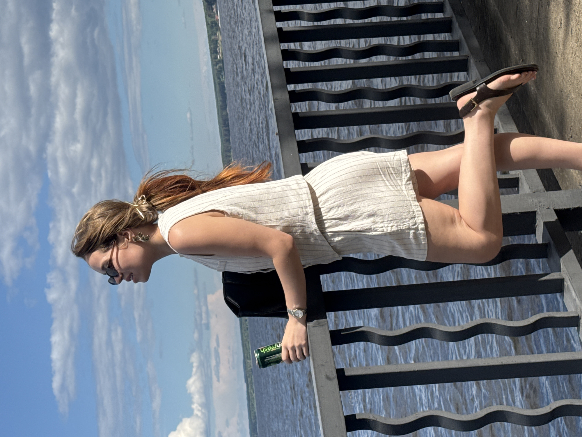 A young woman with auburn hair pulled back in a ponytail, wearing sunglasses and a light-colored striped sleeveless top and shorts, stands on a paved walkway next to a dark metal railing. She holds a green can of beverage in her right hand. Her left leg is bent at the knee and extended backward, with her left foot wearing a dark sandal resting on the walkway. The background features a wide body of water with trees and buildings visible on the far bank, under a partly cloudy blue sky. This location is consistent with the embankment of the Kama River in the city of Chaikovsky, Russia.