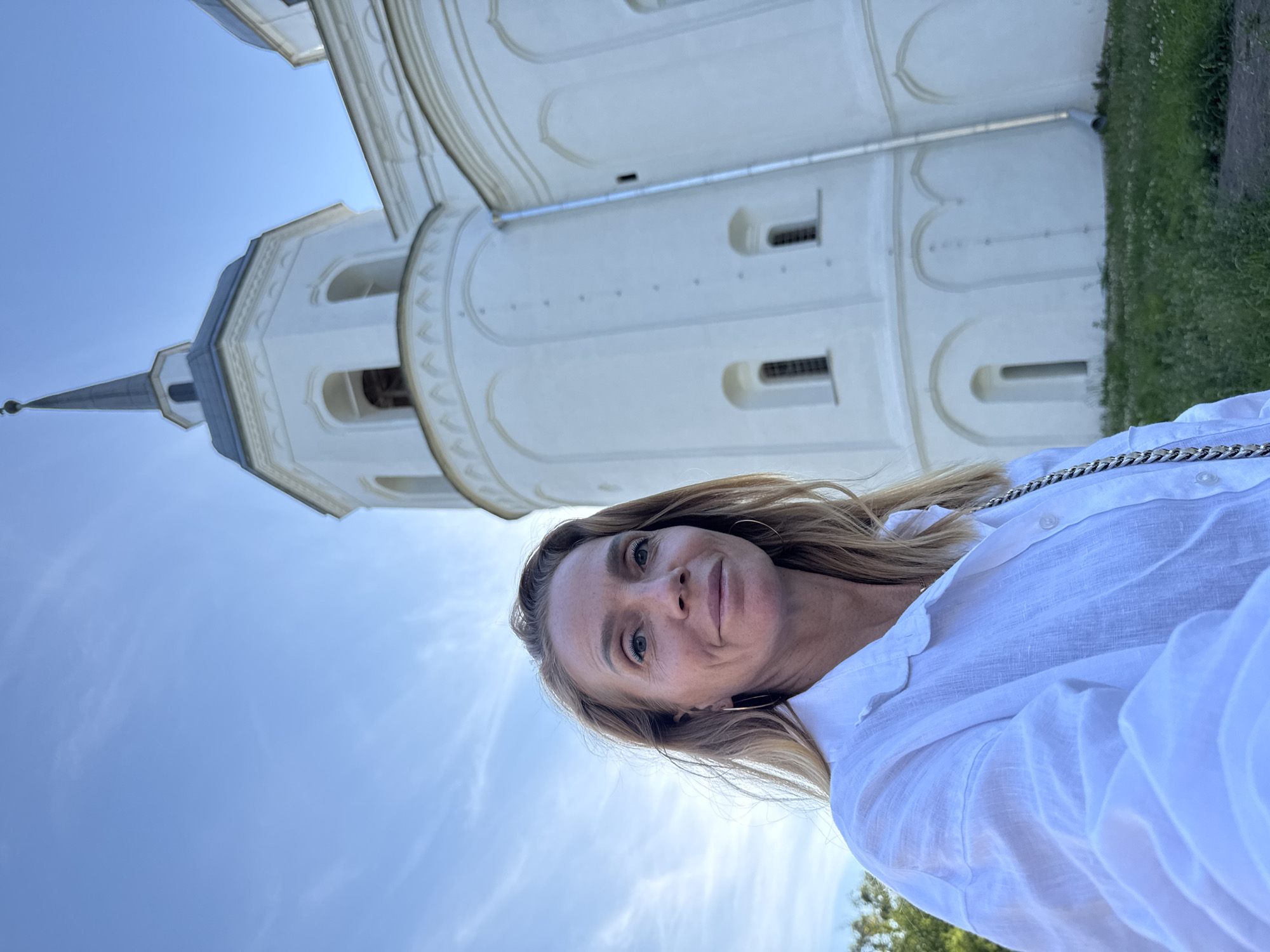 A woman with blonde hair and fair skin is taking a selfie outdoors. She is wearing a white linen shirt and large hoop earrings. Behind her is a tall, white, multi-tiered building with a pointed spire, which appears to be a church or cathedral. The building has arched windows and decorative architectural elements. The sky above is blue with wispy white clouds. Green grass and some foliage are visible in the foreground and at the base of the building. The architecture suggests the location is likely in Veliky Novgorod, Russia, possibly the Saint Sophia Cathedral or a similar historic religious structure.