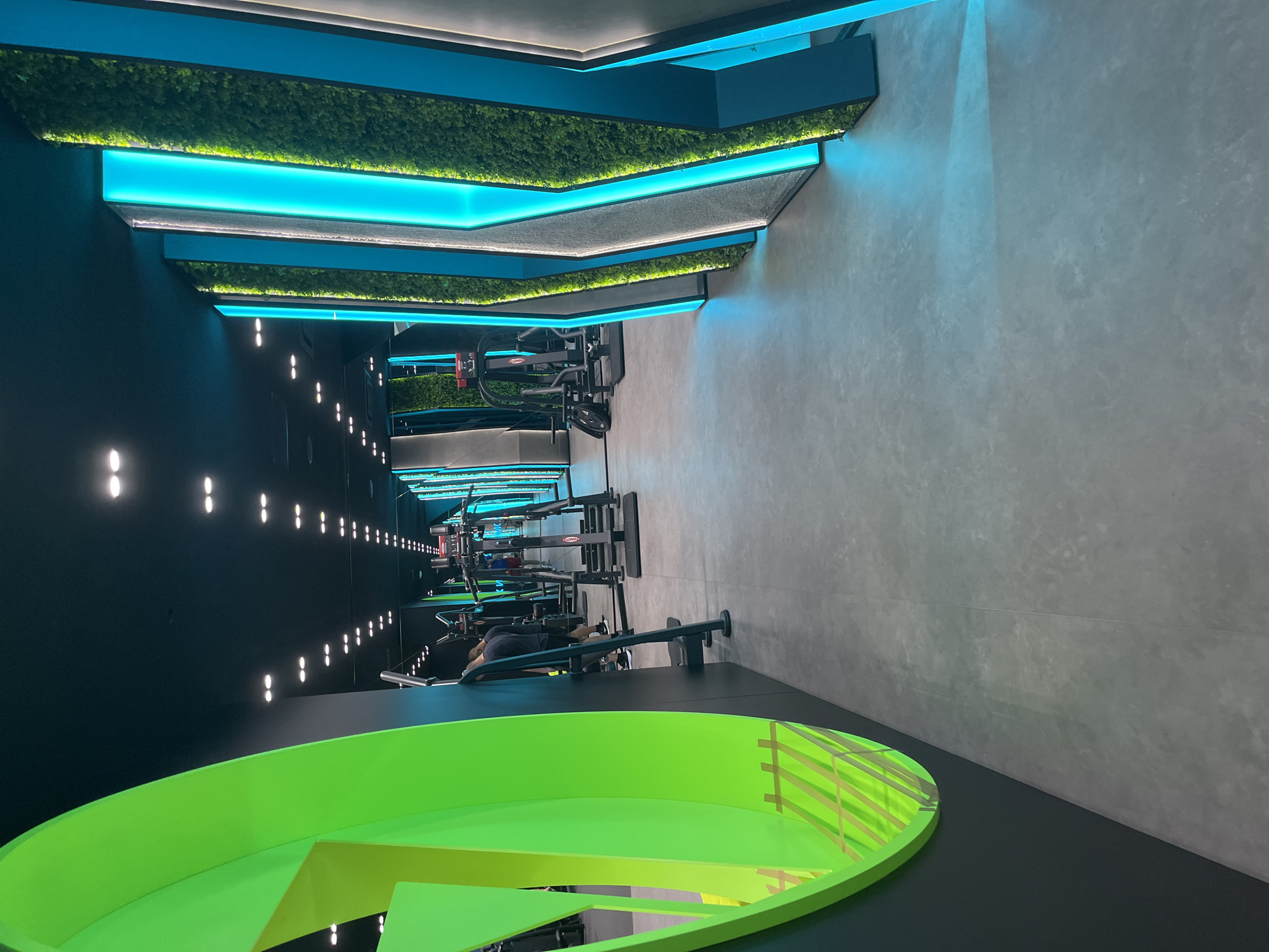 The image captures the interior of a fitness center located in an unknown city. The room is oriented lengthwise, with rows of exercise machines visible. Overhead, the ceiling features a modern design with tiered, angular panels adorned with greenery and illuminated by blue lighting. A prominent green, circular structure with a geometric design is visible in the foreground. One person can be seen using one of the machines. The walls are colored in contrasting black and light grey, with a series of lights running along the black wall.