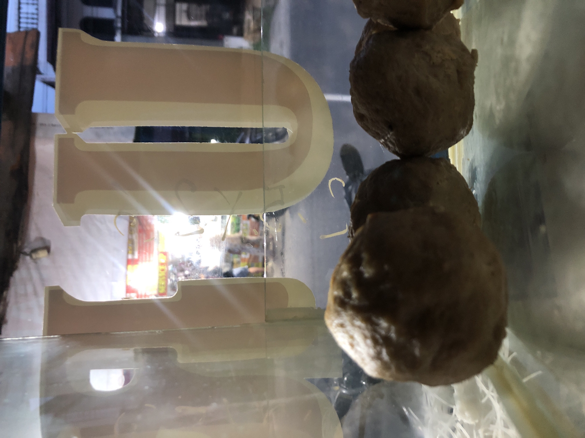 A close-up, low-angle shot captures several large, brown meatballs in the foreground, presented within a glass display case. The meatballs are round and have a textured surface. Behind them, a portion of a building is visible, with what appear to be large, light-colored letters forming an archway. The letters are illuminated, creating a soft glow. In the background, beyond the display case, a street scene unfolds. The street is paved, and there are indications of buildings with windows and architectural details. A few figures, possibly pedestrians or vendors, are visible in the distance, along with a motorcycle. The overall lighting suggests it is either dusk or nighttime, with artificial lights illuminating the scene. The presence of a street and buildings indicates an urban or semi-urban environment.