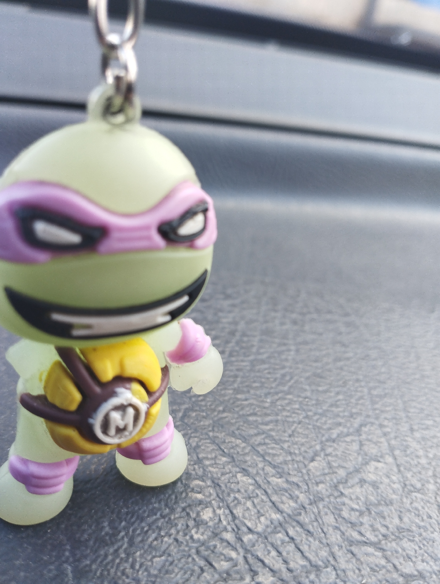 A pale green action figure, resembling a Teenage Mutant Ninja Turtle, is positioned in the center of the frame. The figure is wearing a purple mask over its eyes and a yellow harness with a brown strap across its chest. A circular emblem with the letter "M" is visible on the harness. The figure's hands and feet are a pale translucent green, with pink accents on its knees and elbows. The figure is standing on a dark, textured surface, which appears to be the dashboard of a vehicle. The background is blurred, showing a dark horizontal line and lighter grey tones, suggesting an indoor environment, possibly a car interior. The lighting suggests it is daytime. There is no visible indication of the city of Chaikovsky, Russia, in this image.