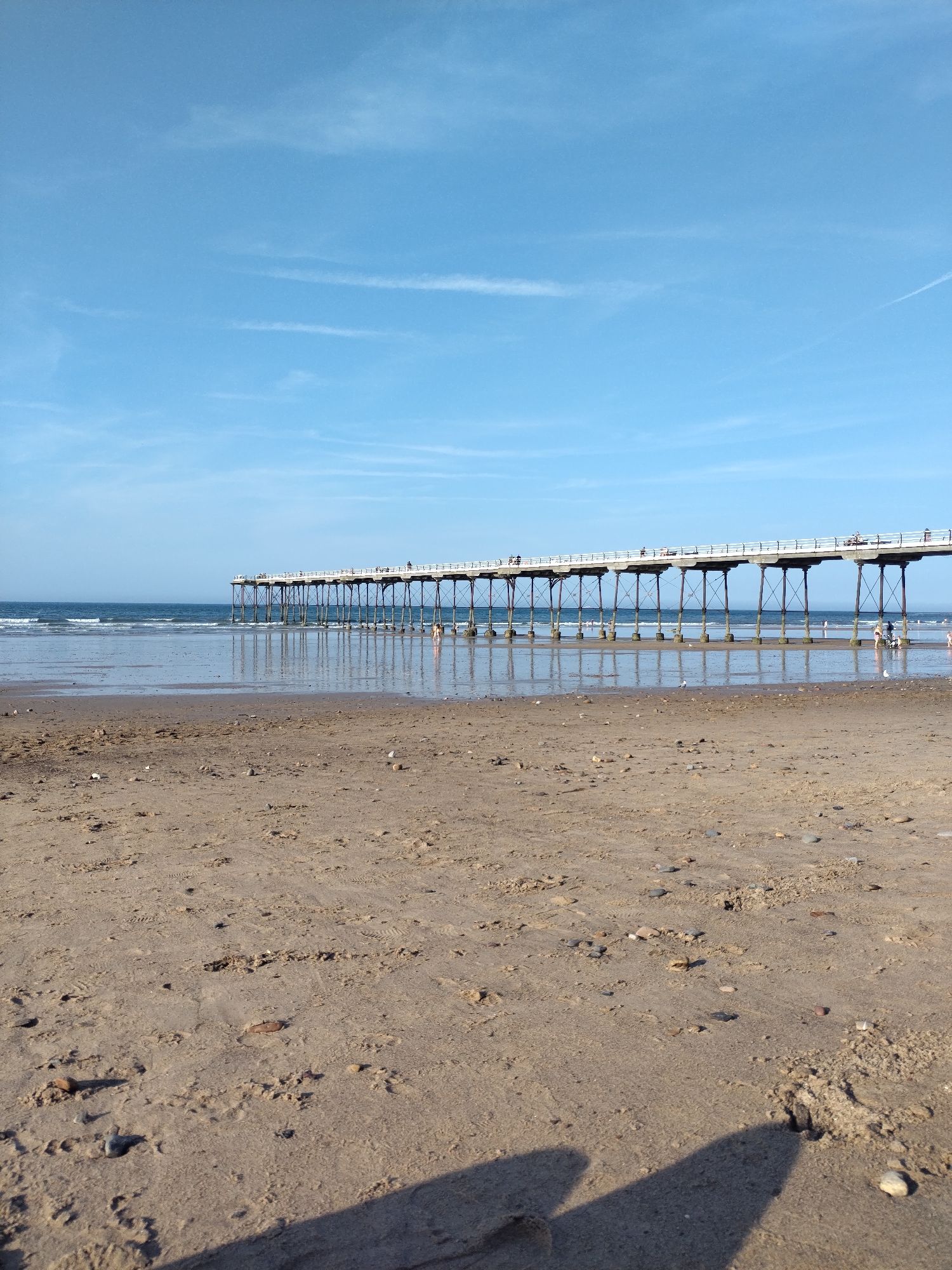 The image shows a view of Saltburn Pier and the beach in Saltburn by the Sea, United Kingdom. The pier, a long, elevated structure with railings, stretches horizontally across the frame, supported by numerous dark, slender pillars. Several indistinct figures are visible walking on the pier. The sea is calm, with gentle waves lapping at the shore. The foreground is a sandy beach with footprints and scattered pebbles. The sky is a clear blue with a few wispy white clouds. The lighting suggests it is daytime, likely in the afternoon, given the shadows cast on the sand.
