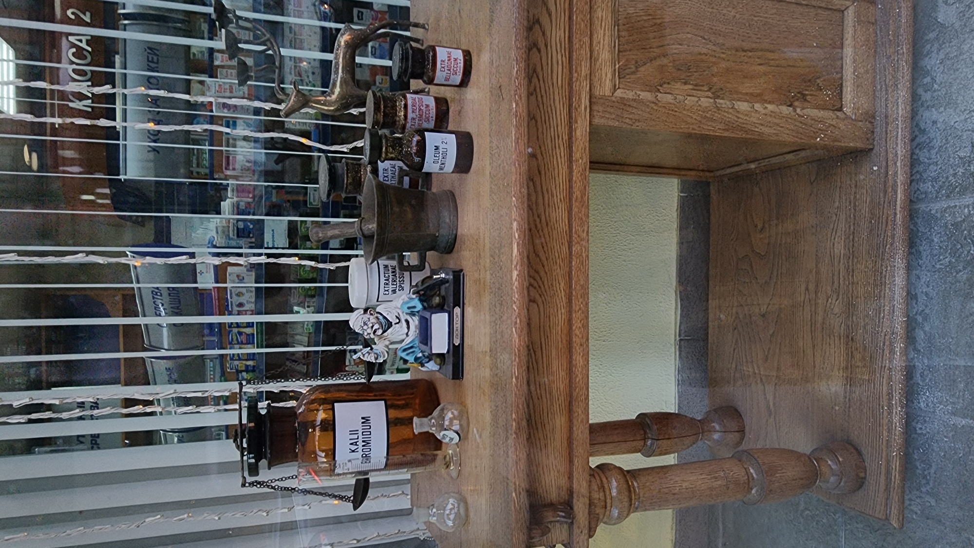 This image shows a display in a shop window, likely a pharmacy or apothecary, in Moscow, Russia. The display sits on a rustic wooden table.
Dominating the table is a large amber glass bottle with a label that reads "KALII BROMIDUM". To its left are two small glass vials. To the right of the amber bottle, a small figurine of a doctor is seated at a desk with a miniature inkwell. Next to the figurine is a white jar with a label that reads "EXTRACTUM VALERIANAE SPISS". A mortar and pestle are positioned behind the white jar. Further to the right are three small brown glass bottles with black caps. The labels on these bottles read: "OLEUM MENTHOLI 2", "EXTR. MERBAPE HERMODFSIDUS SICCUM", and "EXTR. BELLADONNAE SICCUM". In the background, a brass deer figurine stands to the right of the bottles.
The shop window itself has vertical blinds or a similar covering, with decorative string lights hanging in front of the display. Through the window, shelves stocked with various products, likely medications, are visible. Text on the background signage appears to be in Cyrillic, consistent with a Russian location. One sign clearly visible reads "КАССА 2" (KASSA 2), meaning "Cashier 2".
The overall impression is a vintage or old-fashioned apothecary display, intended to evoke a sense of history or traditional medicine.