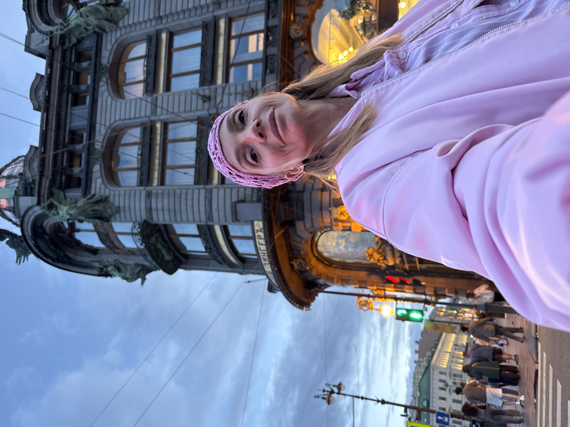 In Saint Petersburg, Russia, a woman in a pink knitted headband and a pink bomber jacket takes a selfie in front of the Singer Building. The building's ornate facade, featuring statues and arched windows, is visible behind her. The sky is overcast and the streetlights are beginning to illuminate the scene. A crosswalk and traffic signals are visible on the street, with a few people walking in the background. The Singer Building, also known as the Dom Knigi (House of Books), is a prominent landmark on Nevsky Prospekt.