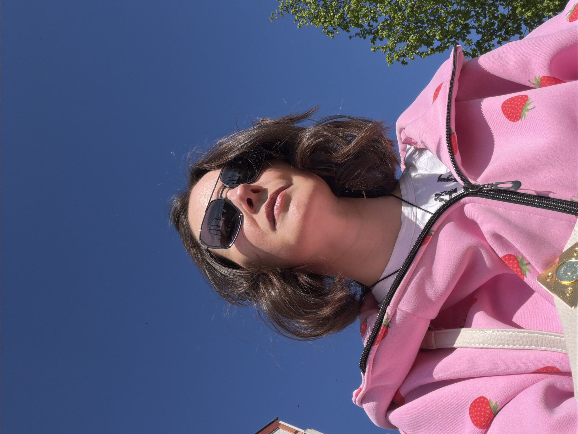 A low-angle shot reveals a person wearing sunglasses and a pink strawberry-patterned hoodie, set against a backdrop of a clear blue sky. Some green foliage is visible in the upper right corner of the frame. At the bottom of the image, a partial view of a building with a red roof can be seen. The location is Unknown, Unknown.