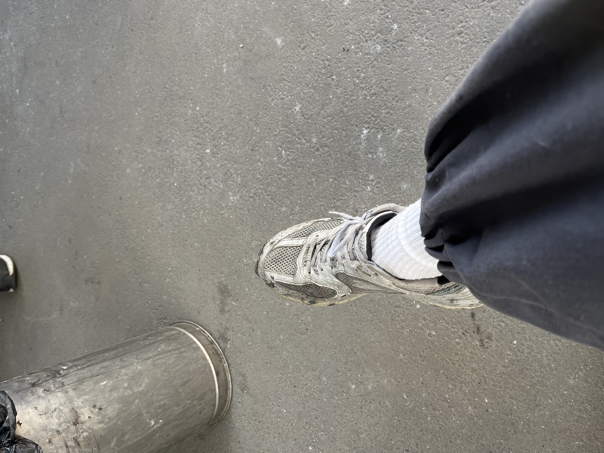 The image depicts a person's leg wearing a grey athletic shoe and white socks, standing on a paved surface next to a cylindrical metal trash can. The trash can is positioned to the left of the frame and appears to be made of brushed metal. The ground is a grey, speckled pavement with some scattered debris. The lighting suggests an outdoor environment, possibly in the daytime. There is no specific indication of the location being Kyiv, Ukraine, within the image itself; this information is external context.