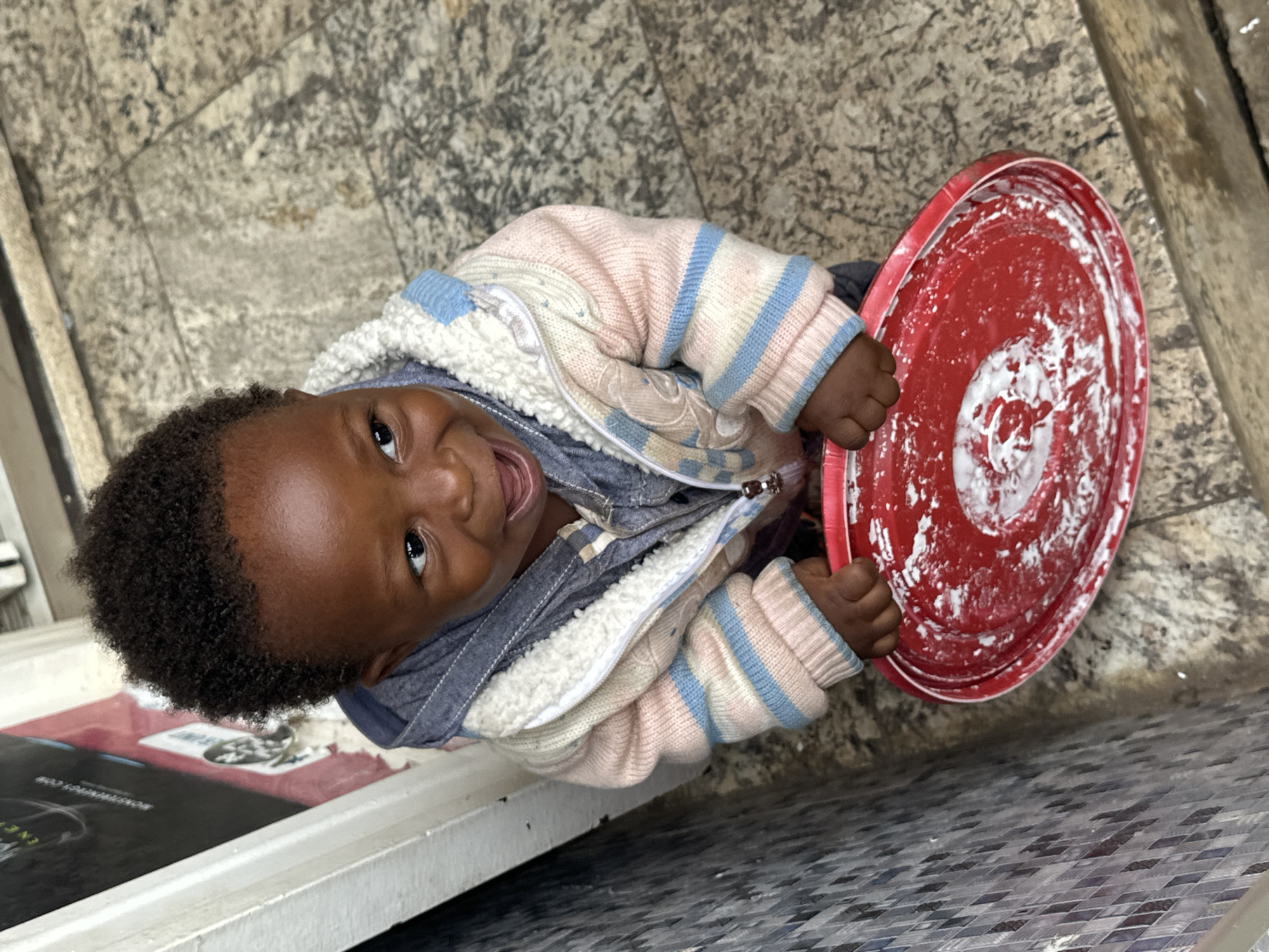 A high-angle, full shot captures a young child smiling at the camera, holding a red plastic bowl with white residue on its surface. The child is wearing a pink and blue striped sweater over a denim jacket, with a white furry lining visible on the jacket. The child's skin is dark, and their curly hair is dark brown. The child's eyes are wide open, and their mouth is open in a smile. The child is positioned in the center of the frame.
The background consists of a textured, light-colored floor with a diamond-patterned tile section in the lower right corner. The floor appears to be outdoors, possibly a courtyard or street in Gura Top, Nigeria. The lighting suggests it is daytime. The overall mood is cheerful and innocent, conveyed by the child's bright smile. No visible text is present in the image.