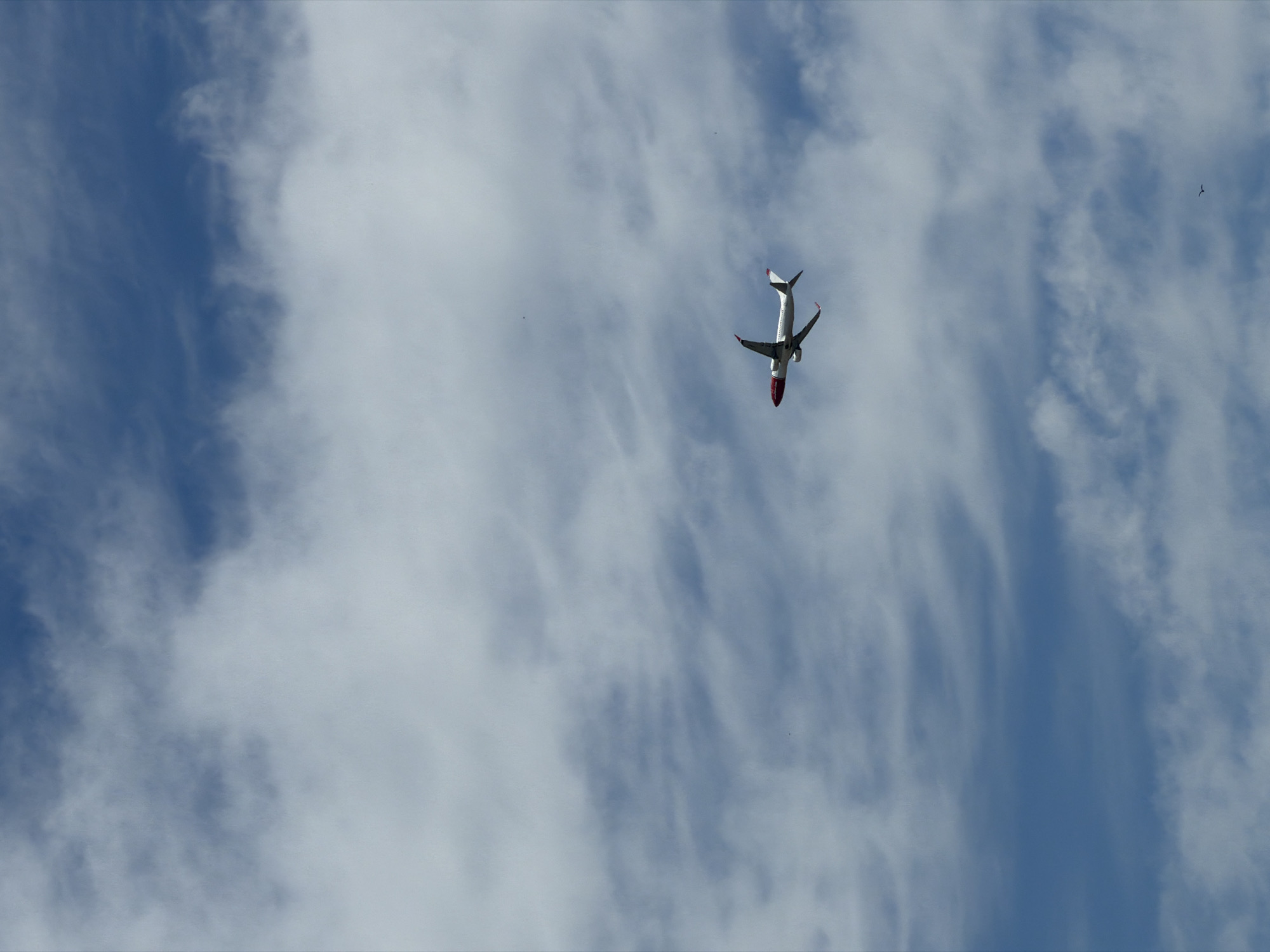 An airplane with a white body and a red nose and tail section flies across a blue sky filled with scattered white clouds. The airplane is in the right-middle portion of the image, angled slightly upwards. A small bird is visible in the lower right corner of the image.