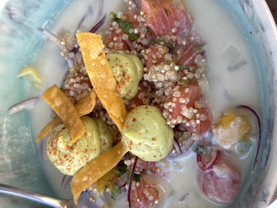 The image displays a close-up, overhead view of a prepared food dish presented in a light blue ceramic bowl with a textured, slightly uneven rim. The dish features a creamy, light green-tinted liquid base, likely a coconut milk or leche de tigre. Submerged and partially submerged ingredients include: diced pinkish-red segments, consistent with fish or seafood; numerous small, white, translucent grains resembling quinoa; thinly sliced red onions; and small pieces of diced green and yellow vegetables.
Three distinct dollops of a pale green, smooth purée, possibly an avocado or herb cream, are strategically placed on top of the solid ingredients, each lightly sprinkled with reddish-orange spice flakes. Several crisp, golden-yellow, elongated strips, resembling fried wonton or tortilla strips, are arranged across the dish, intersecting the green purée dollops. A metallic spoon handle is visible along the upper left edge of the bowl, partially submerged in the liquid.
The scene is a still life of a culinary presentation, likely captured in a restaurant or dining setting in Aldoar, Foz do Douro e Nevogilde, Portugal. No other people, actions, or specific environmental details beyond the dish and bowl are discernible.