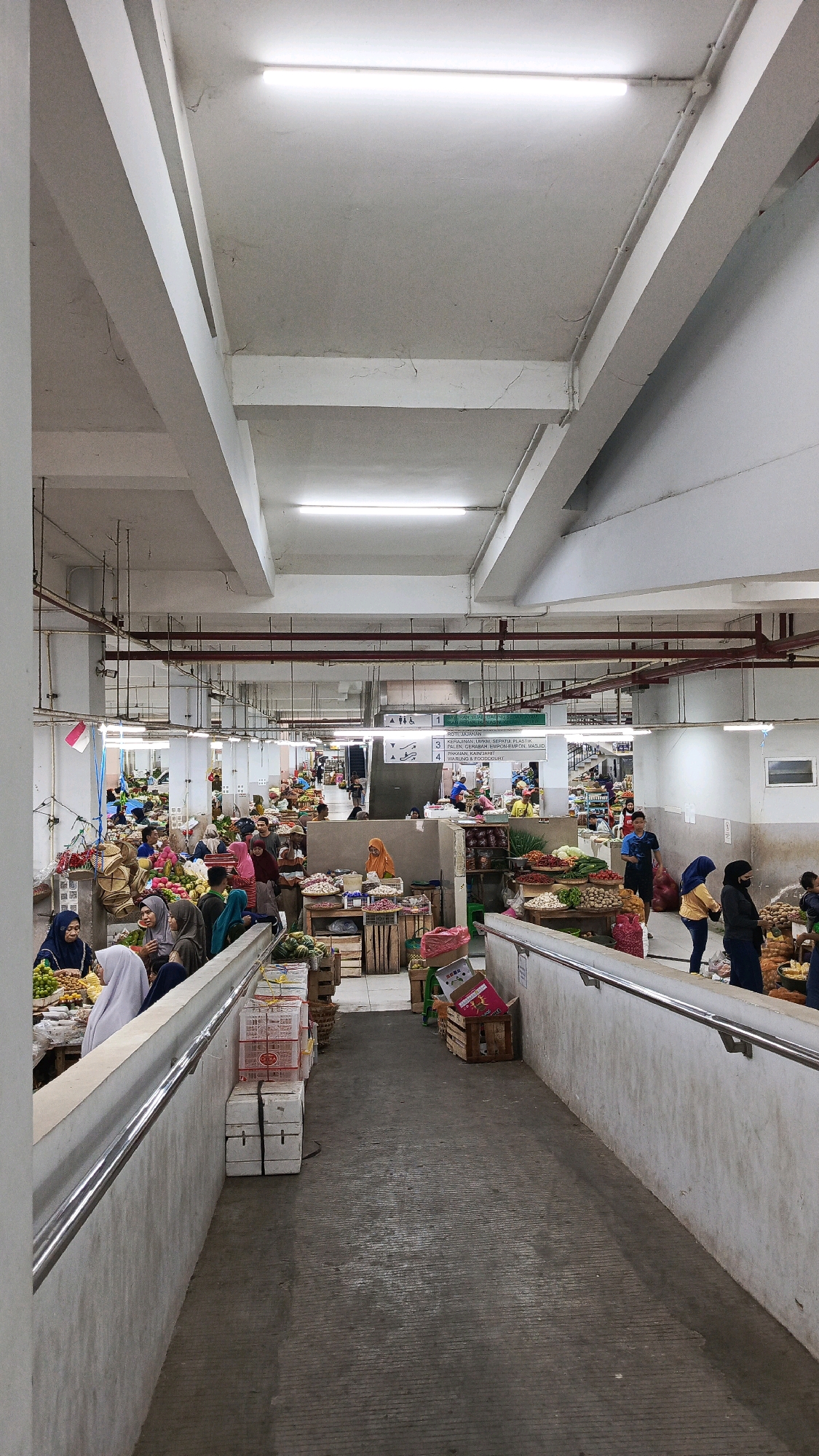 The image depicts the interior of a bustling indoor market, identified as being in Nologaten, Indonesia. The market is characterized by stalls selling a variety of produce, including fruits and vegetables, displayed in crates and on tables. Several individuals are present, engaged in shopping or selling activities. Most shoppers are women, many wearing headscarves, and some are seen interacting with vendors or examining the goods. The environment is utilitarian, with concrete floors and ramps, exposed ceiling beams, and fluorescent lighting. Baskets, boxes, and various market paraphernalia are visible throughout the scene, contributing to the busy atmosphere of a commercial hub.