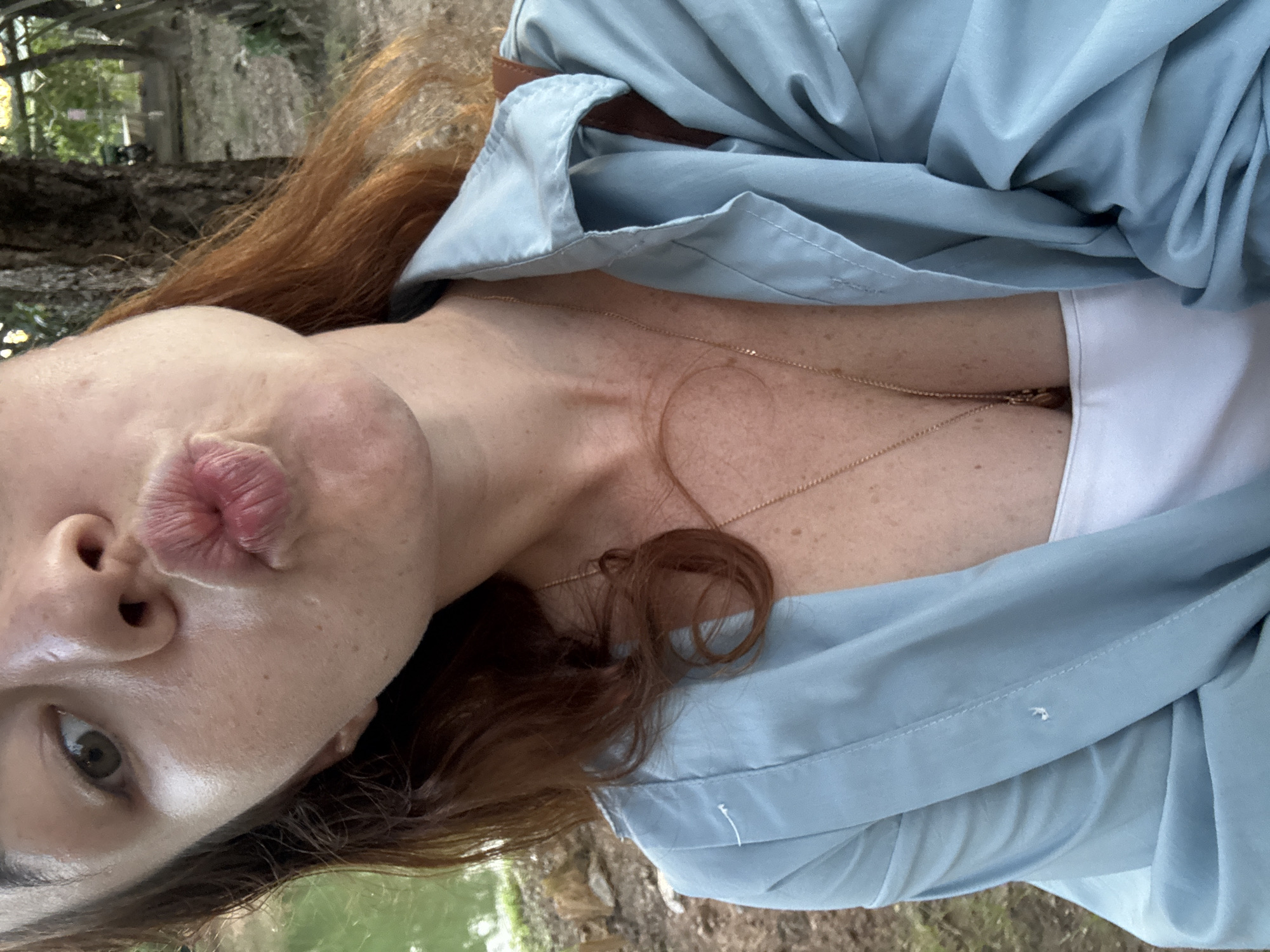This is a close-up, vertically oriented selfie of a person with auburn hair, outdoors. The subject is leaning in, puckering their lips for a kiss. Their face is partially visible, with one eye looking towards the camera. The person is wearing a light blue, open collared shirt over a white top, and a delicate gold necklace. The background appears to be a natural setting with trees and possibly water, suggesting a park or garden. The lighting indicates it is daytime, possibly morning or afternoon, with soft, natural light. There is no visible text in the image. The overall mood is playful and intimate.
