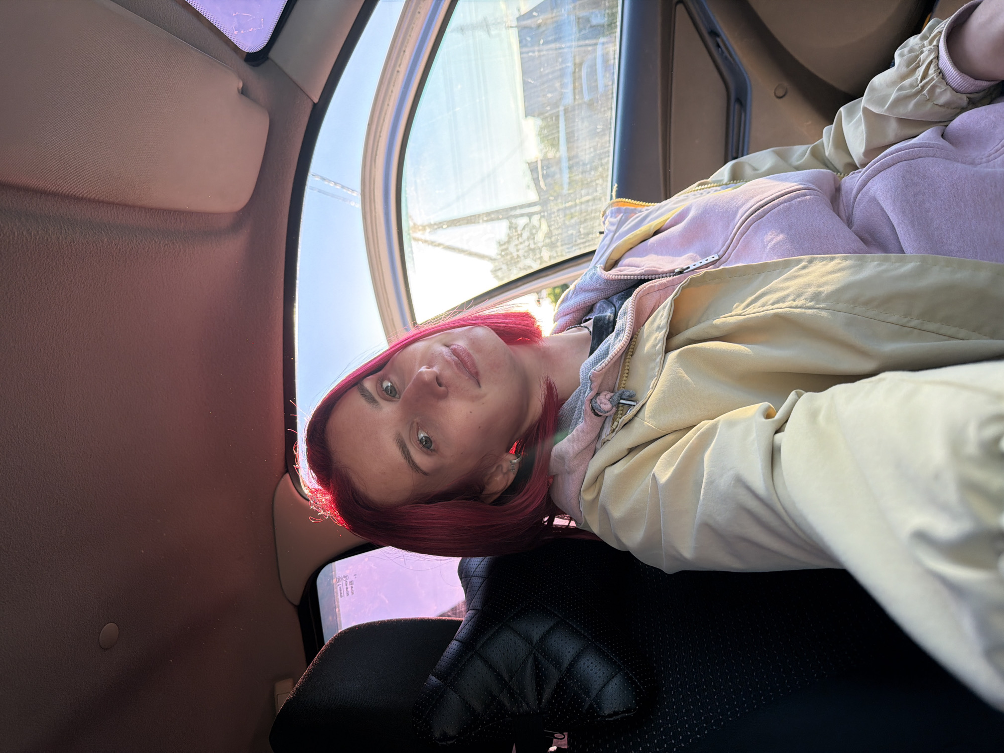 The primary subject is a young woman with striking bright pink hair, captured in a close-up selfie or portrait. She is wearing a light pink zippered hoodie layered under a light beige or khaki-colored jacket. The image is rotated 90 degrees clockwise, positioning her head on the left and her body extending to the right.
The setting is the interior of a car, specifically the passenger side. Visible car elements include a beige sun visor, a car door frame, a window through which exterior light and faint reflections are seen, and a black car seat with a distinctive quilted pattern on the headrest.
The woman appears to be taking a photograph of herself while seated in the vehicle, displaying a relaxed demeanor and a faint smile.
Notable details indicate a clear, sunny day, with bright natural light streaming in through the window, illuminating the scene and creating a strong highlight on her pink hair. Through the window, indistinct external structures, possibly buildings or power lines, are faintly visible. Faint, undecipherable text or numbers can be seen on the car window, which may be reflections or decals. The overall atmosphere is casual and vibrant due to the bright lighting and the subject's colorful hair. The location context is Chaikovsky, Russia.