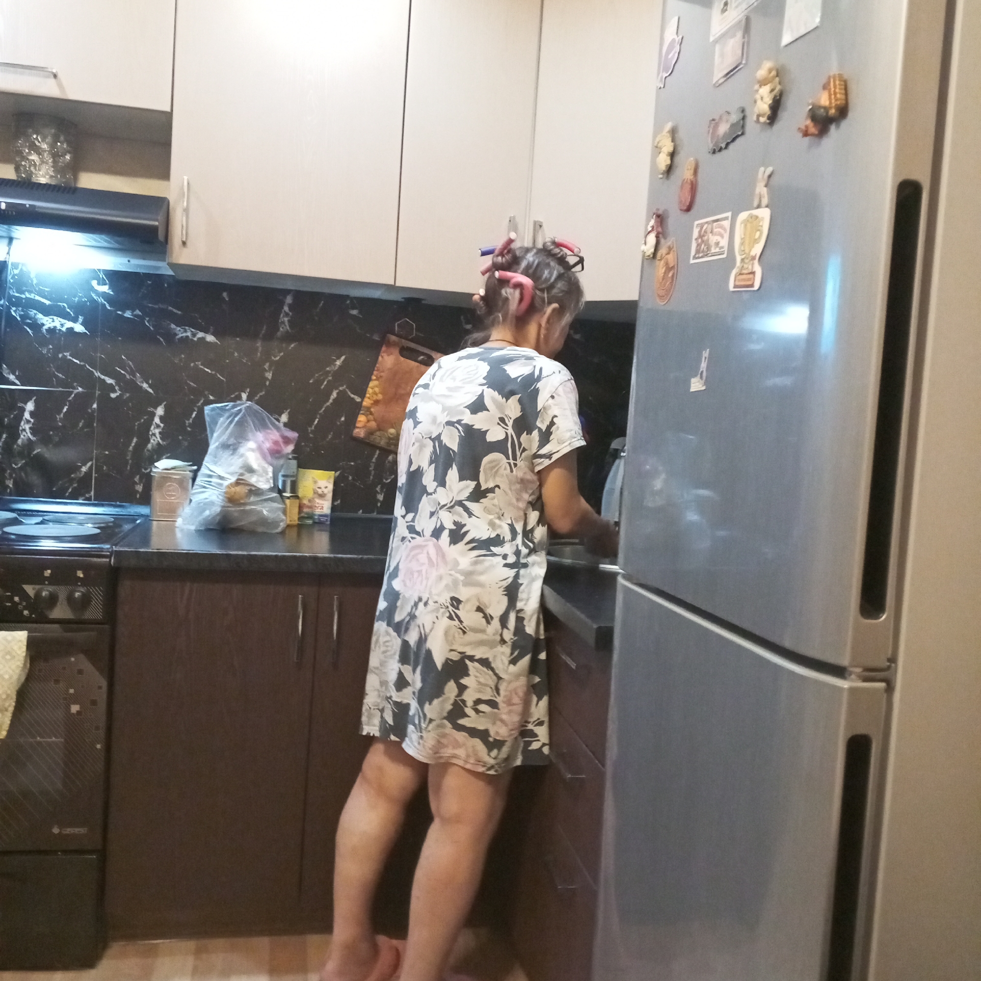 The image shows a woman in a kitchen, possibly in Prokopyevsk, Russia. She is standing near a refrigerator, which has magnets on it. The woman is wearing a floral dress and has hair rollers in her hair. There is a stove and cabinets in the background. The kitchen appears to be a typical home kitchen.