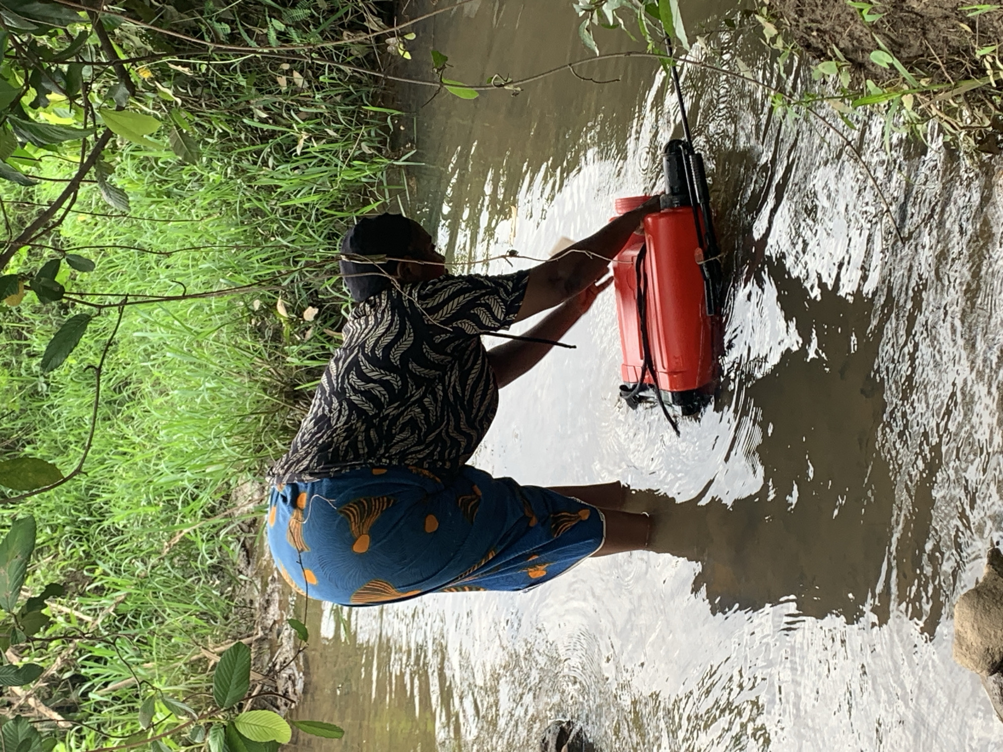 The image shows a person in a stream in Agban, Nigeria. The individual is crouched in the water wearing a dark patterned shirt and blue patterned shorts. They are interacting with a large, orange object that appears to be a container or piece of equipment partially submerged in the stream. The surrounding environment includes lush green vegetation on the banks of the stream, with trees and grass visible. The water is murky.