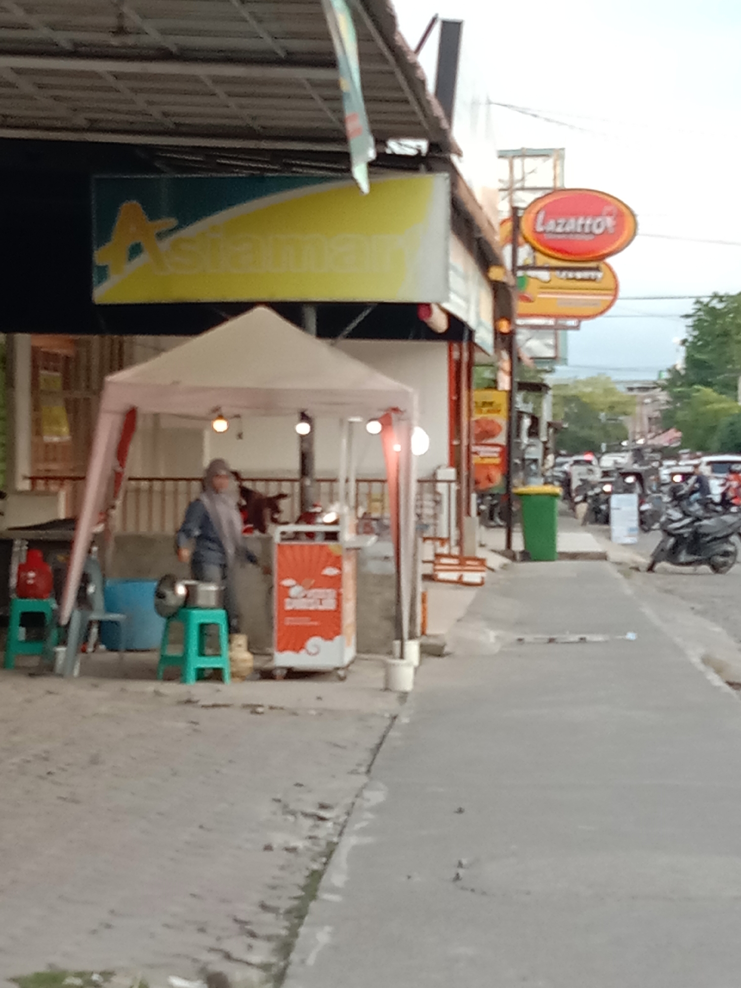 The scene captures a street view in Lhokseumawe, Indonesia. A vendor is operating a small food stall set up on the sidewalk next to a building with an "Asiaman" sign. The vendor, wearing a headscarf, is standing near a cart with an orange design, appearing to be preparing food. In the background, traffic is visible, including motorcycles and cars. A green trash can is present along the sidewalk. The "Lazatto" sign is displayed in the distance.