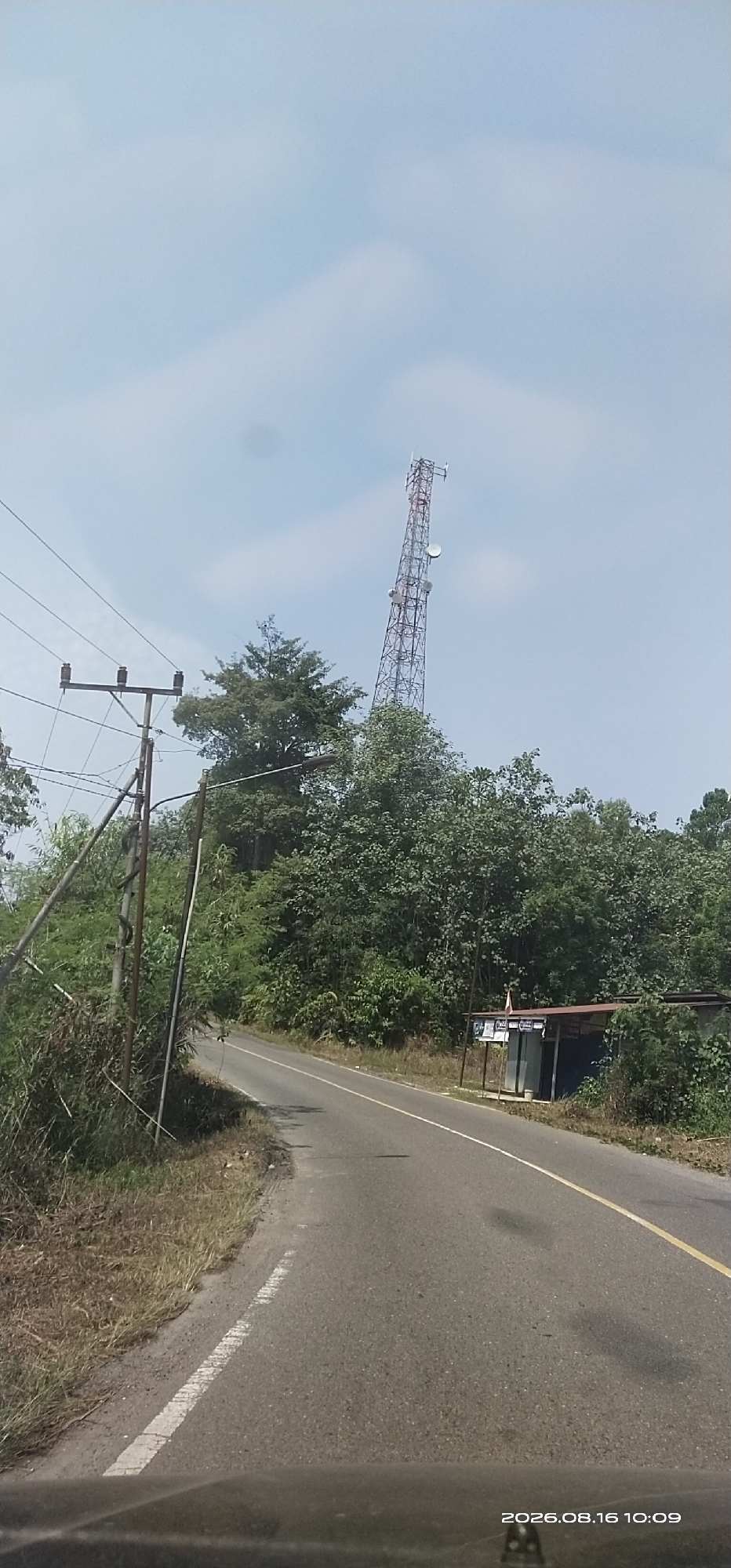 The image captures a view from a vehicle driving on a paved road in Lenggang, Indonesia. The road curves to the left, flanked by lush green vegetation on both sides. In the distance, a tall telecommunications tower rises against a bright, hazy sky, its lattice structure clearly visible. On the right side of the road, a small, open-air kiosk or stall with a corrugated metal roof is situated amongst the trees. Utility poles with multiple insulators and wires stand on the left side of the road, suggesting the presence of electrical infrastructure. The overall scene depicts a rural or semi-rural environment with a blend of natural elements and man-made structures. The timestamp in the lower right corner indicates the image was taken on August 16, 2026, at 10:09 AM.