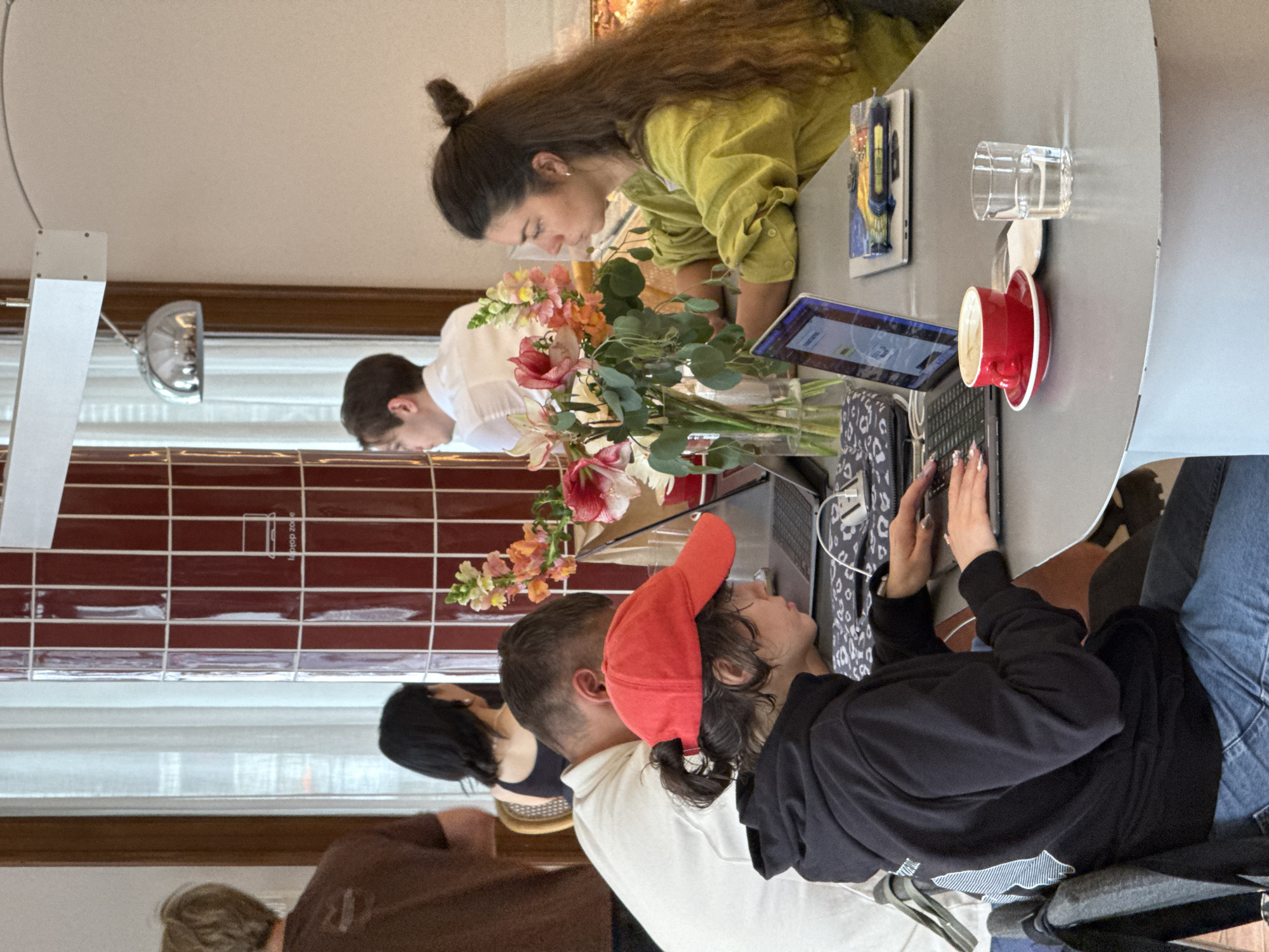 The image captures a shared workspace in Belgrade, Serbia. Four individuals are seated around a grey oval table, each engaged in personal work. One woman with long brown hair, styled in a bun, is wearing a yellow-green shirt and looking at a tablet. Another person, wearing a white shirt, is focused on something out of view. A person wearing a black hoodie and a red baseball cap is actively typing on a laptop. Nearby them, another person in a brown shirt is seated. A vase of flowers sits in the middle of the table, next to a red coffee cup and a glass of water. Natural light enters through a window behind the table.