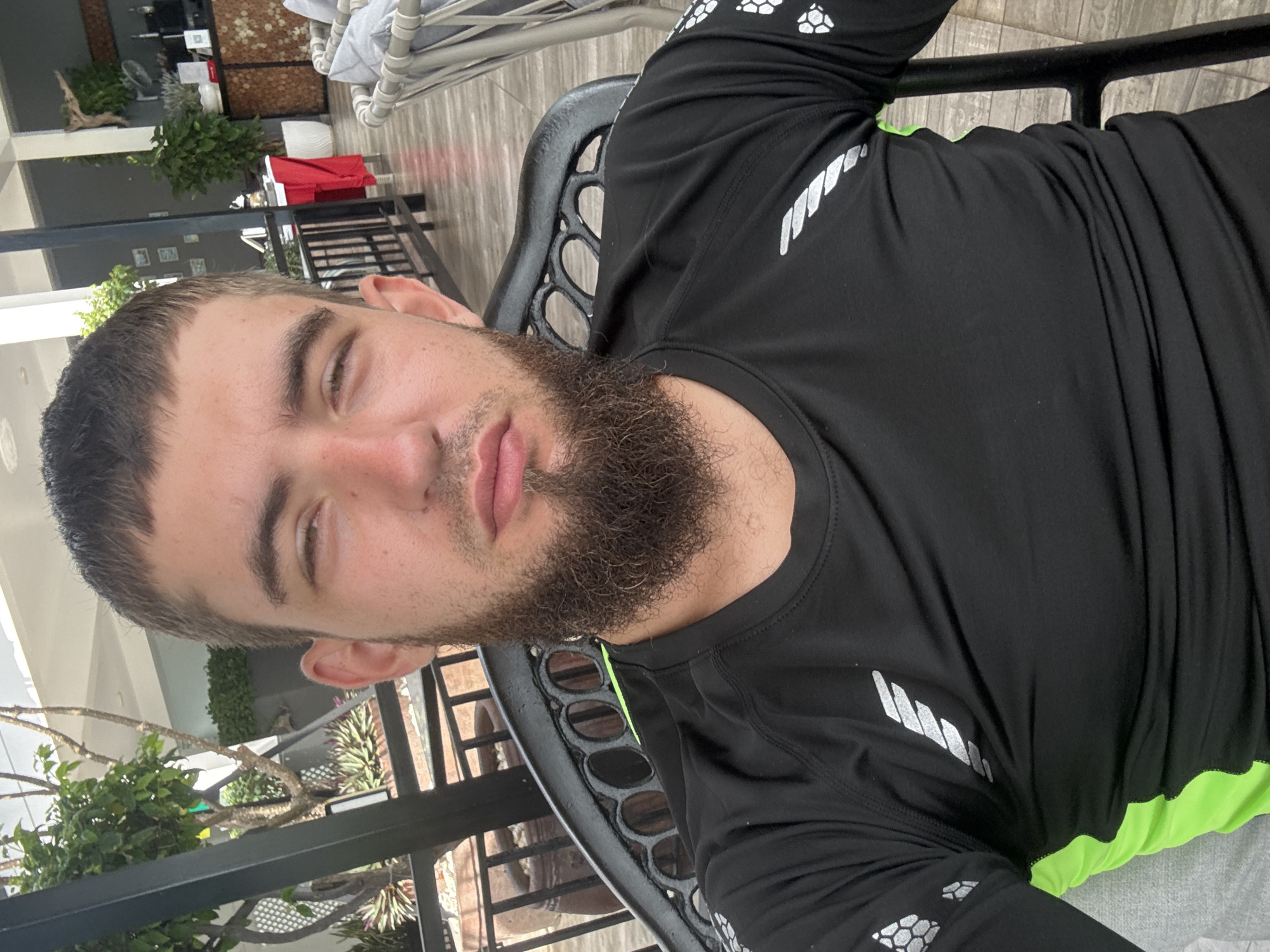 A man with short, dark hair and a full beard is seated in a black chair. He is wearing a black long-sleeved shirt with silver accents on the sleeves and a bright green lining visible at the collar and down the sides. The background suggests an outdoor seating area, possibly a cafe or restaurant, with plants, potted trees, and other seating visible. The flooring is tiled. The man is looking towards the camera with a neutral expression. The overall impression is a casual, daytime setting. The location is identified as Bắc Nha Trang, Vietnam.