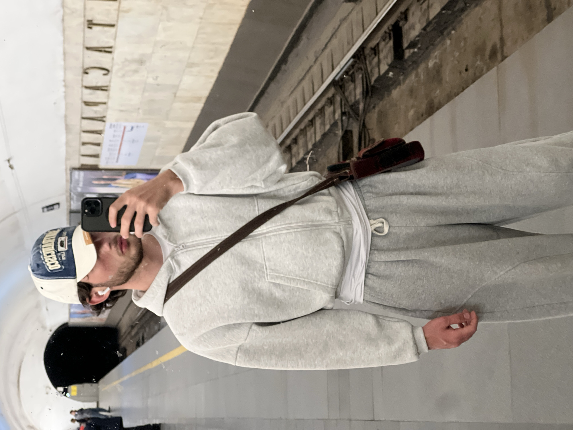 A man in a grey sweatsuit and a baseball cap is taking a selfie in the Yerevan Metro. The reflection in the glass of the station shows the tiled walls and platform. A brown leather strap crosses his body, and a small bag is visible on his upper back. The station walls feature gold lettering. A tunnel entrance is seen behind him.