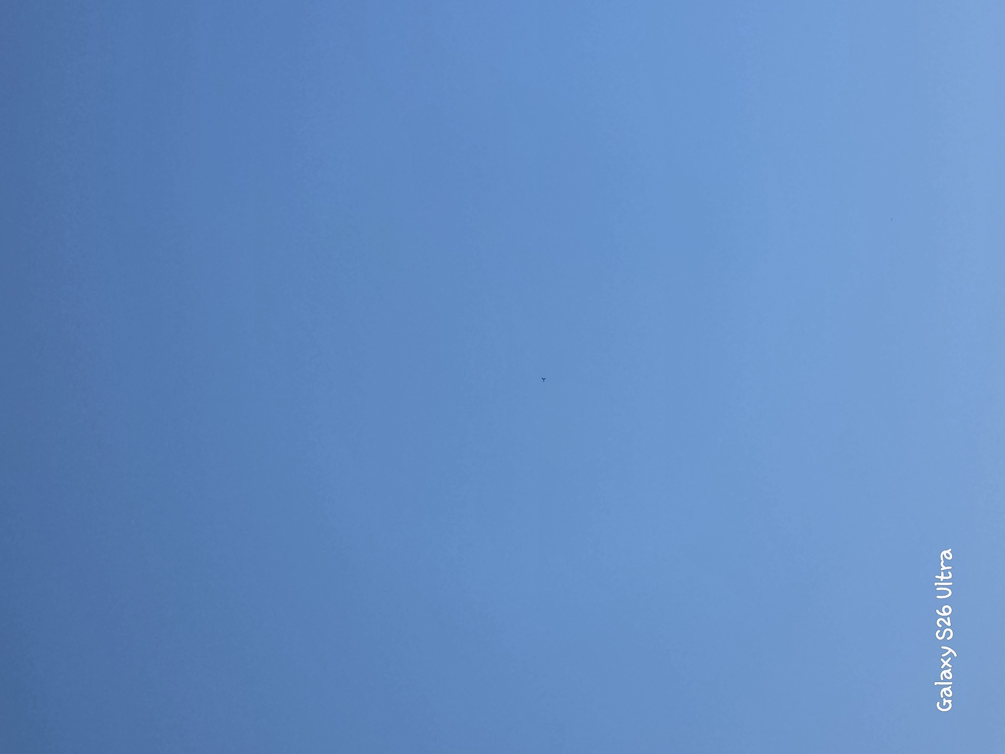 A clear, bright blue sky dominates the image, with no clouds visible. A small, dark, distant object that appears to be an airplane or a bird is captured against the sky in the upper-middle portion of the frame. In the lower-left corner, the text "Galaxy S26 Ultra" is watermarked in white, handwritten font. The scene is devoid of any specific Plovdiv landmarks or any indication of the city.