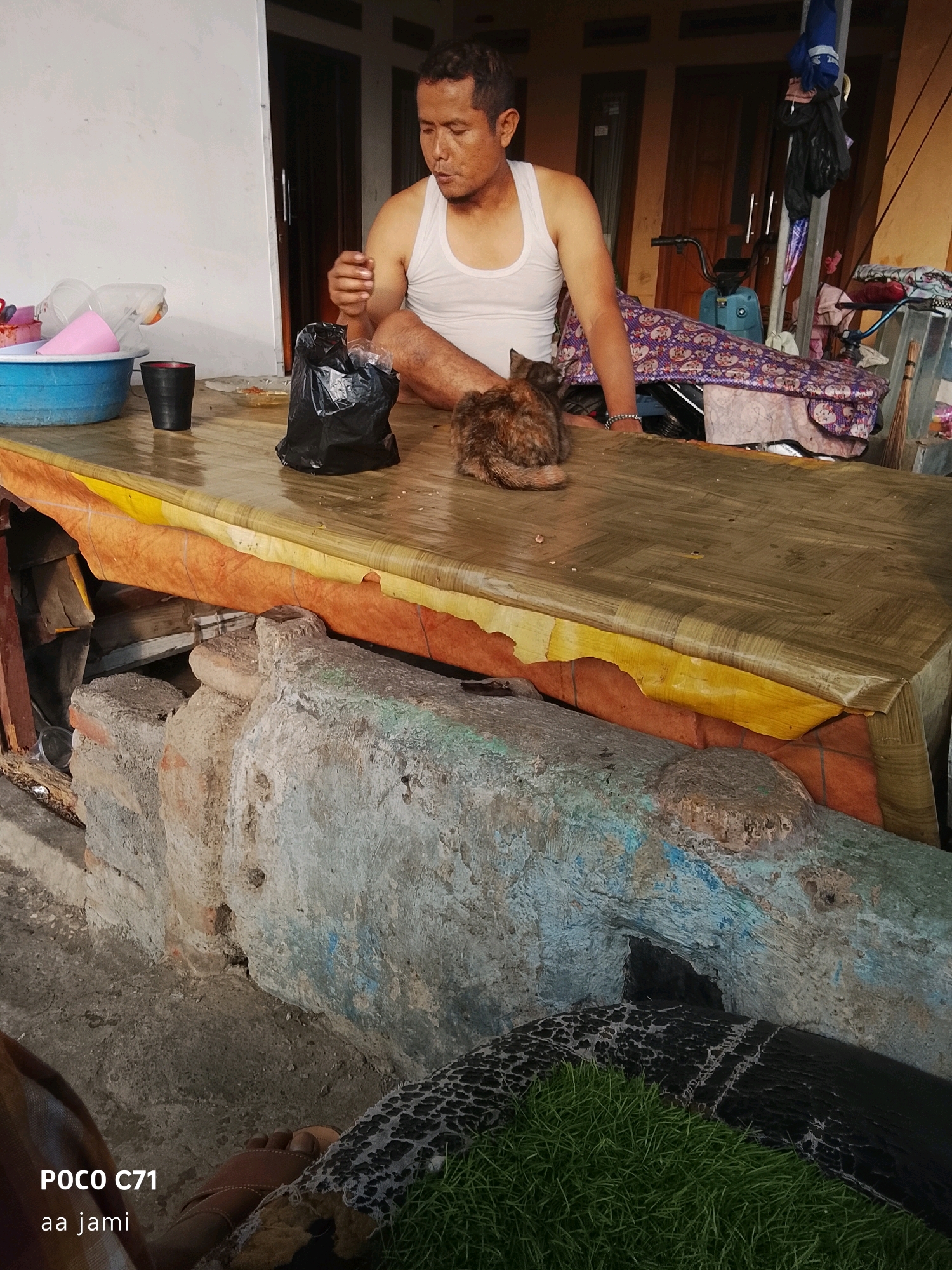 A man wearing a white tank top sits on a table in what appears to be an outdoor or semi-outdoor setting. He is looking down at a small, tabby cat resting beside him on the table. A black plastic bag is placed on the table near the man. In the background, a blue motor scooter is visible, along with various items that suggest a home environment, possibly in a rural or suburban area of Tasikmalaya, Indonesia. The table is covered with a patterned vinyl cloth, and a rough, textured surface, possibly a stone or concrete structure, supports the table. The ground in front of the table is uneven and appears to be made of concrete or packed dirt.