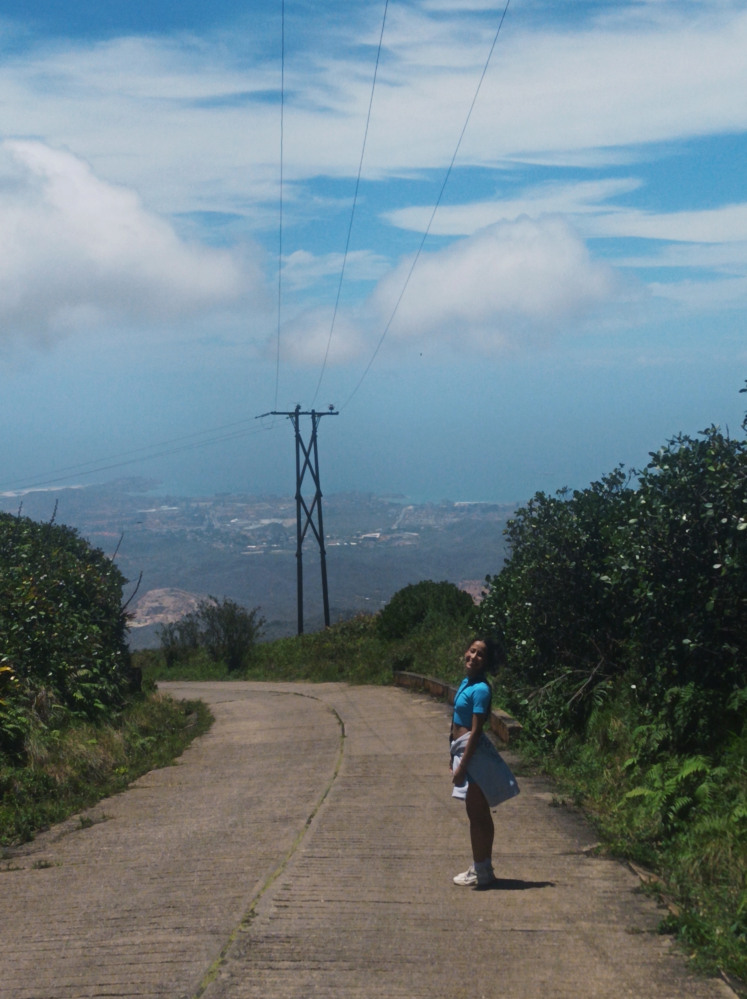 The image shows a young woman standing on a paved road that curves towards the background. She is wearing a blue athletic top, shorts, and white sneakers, and has a white jacket tied around her waist. She is smiling and looking towards the camera.
Behind her and down the road is a power line tower, with wires stretching upwards towards the sky. In the distance is a hazy view of what appears to be a city.
The road is lined with dense green foliage on both sides. The sky is blue with scattered white clouds.
Based on the information given, the location is in the city of Las Piedras, Venezuela. The photo appears to be taken at a high elevation, likely a mountain road overlooking the city.